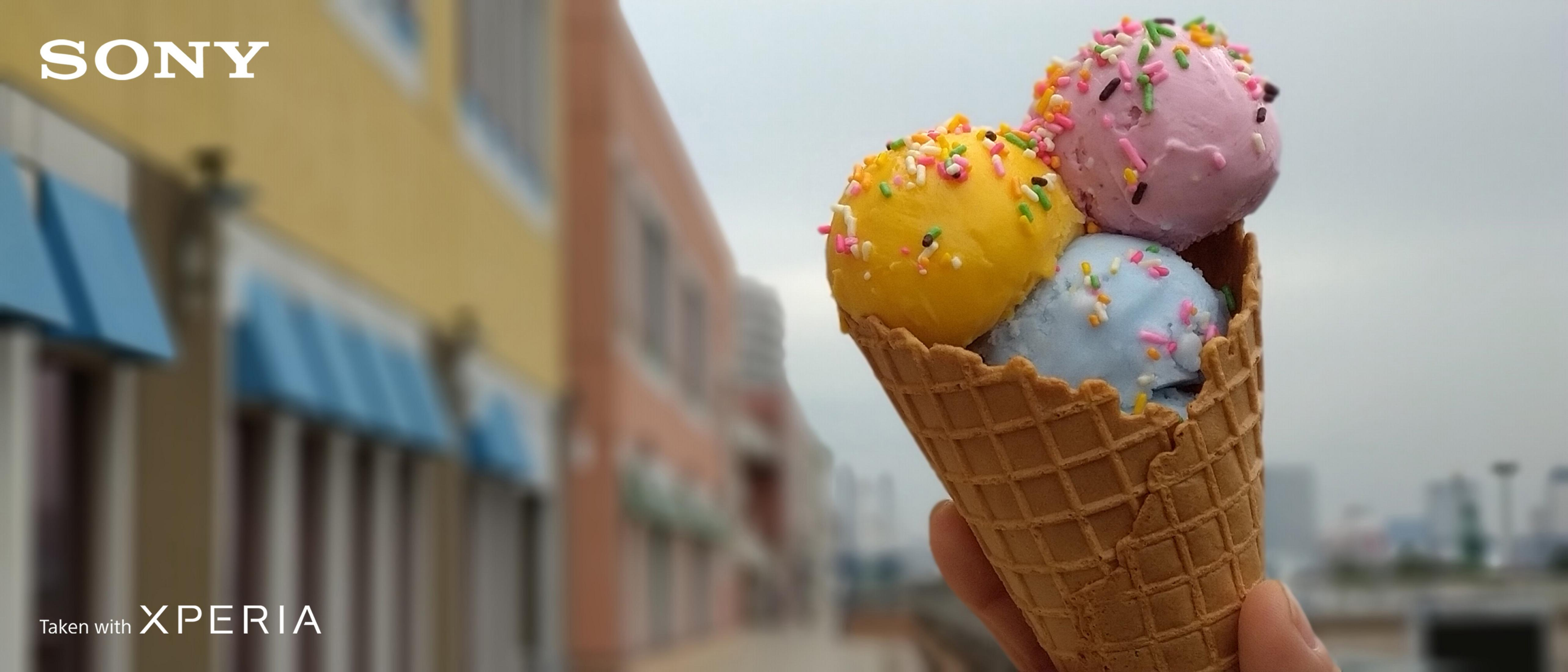 Wafelek z lodami z trzema kulkami, żółtą, różową i niebieską, na tle rozmytej scenerii.