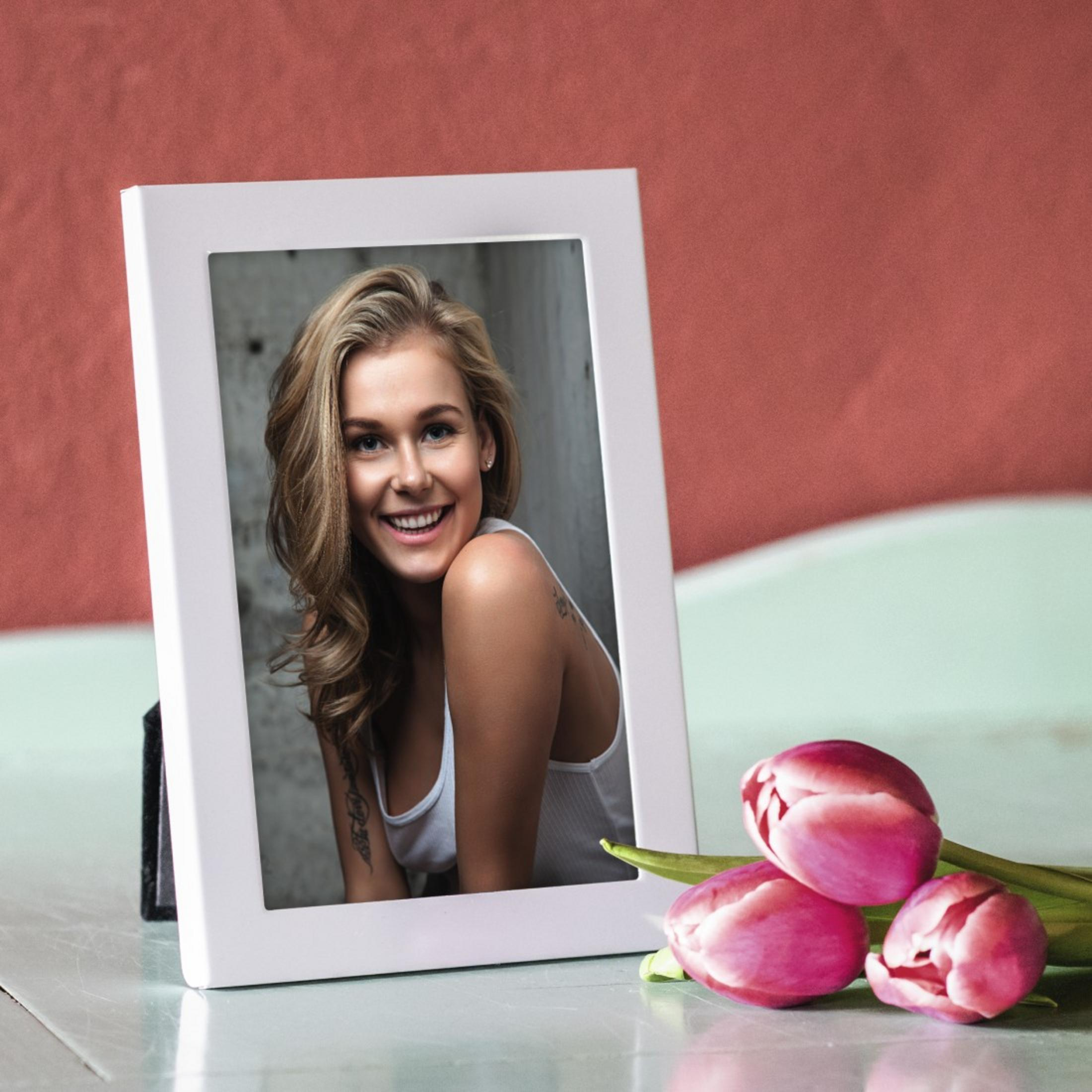 Ein weißer Fotorahmen mit einem Foto einer lächelnden Frau, auf einem Tisch mit drei rosa Tulpen.