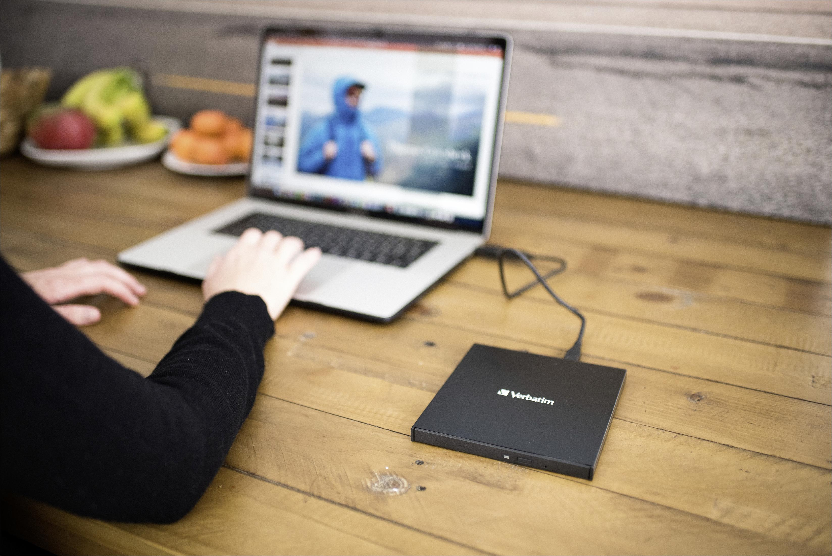 Osoba korzystająca z laptopa z zewnętrznym dyskiem Verbatim na drewnianym stole, z owocami w tle.
