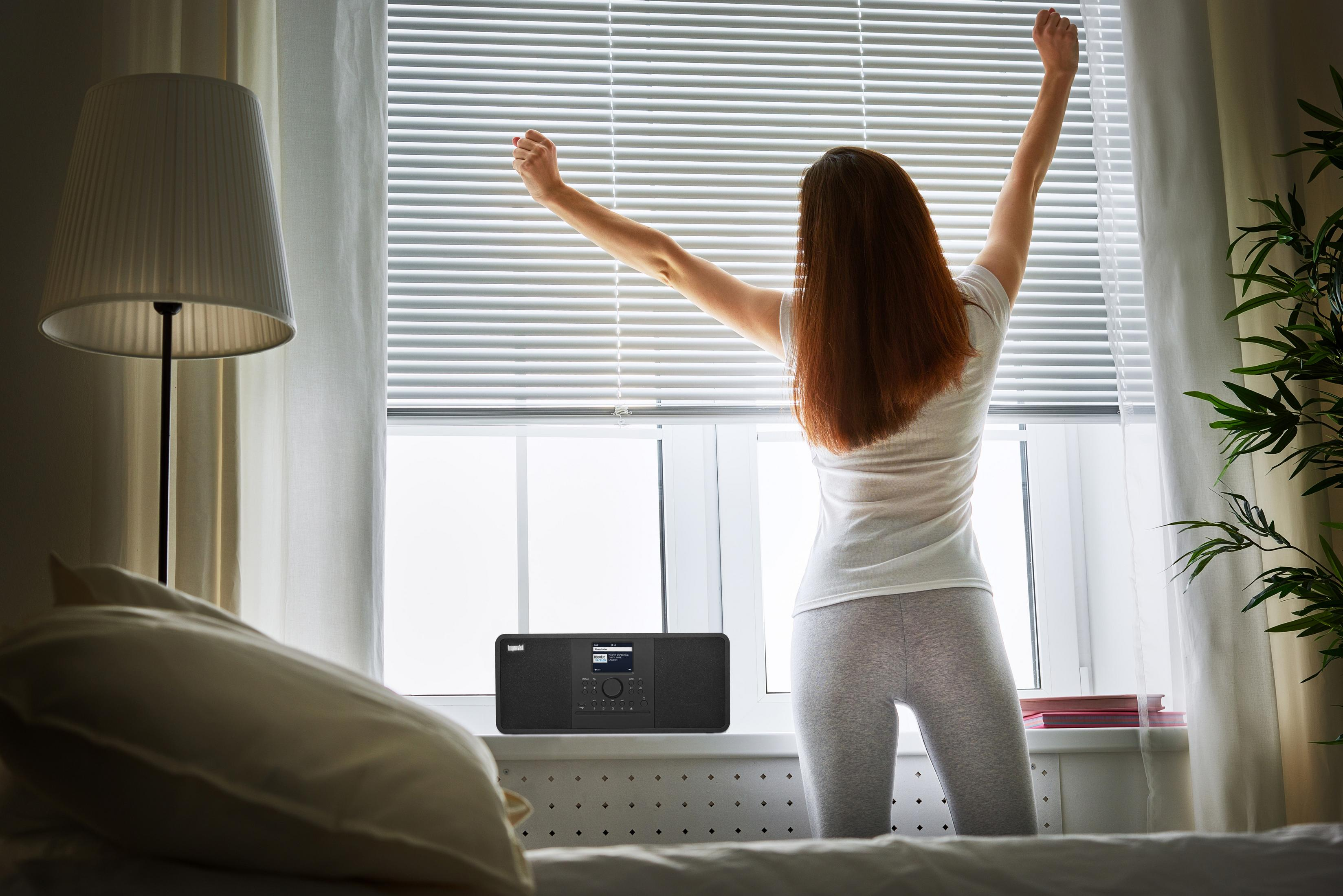 Kobieta wyciąga ręce przy oknie z radiem. Lampa i łóżko na pierwszym planie.