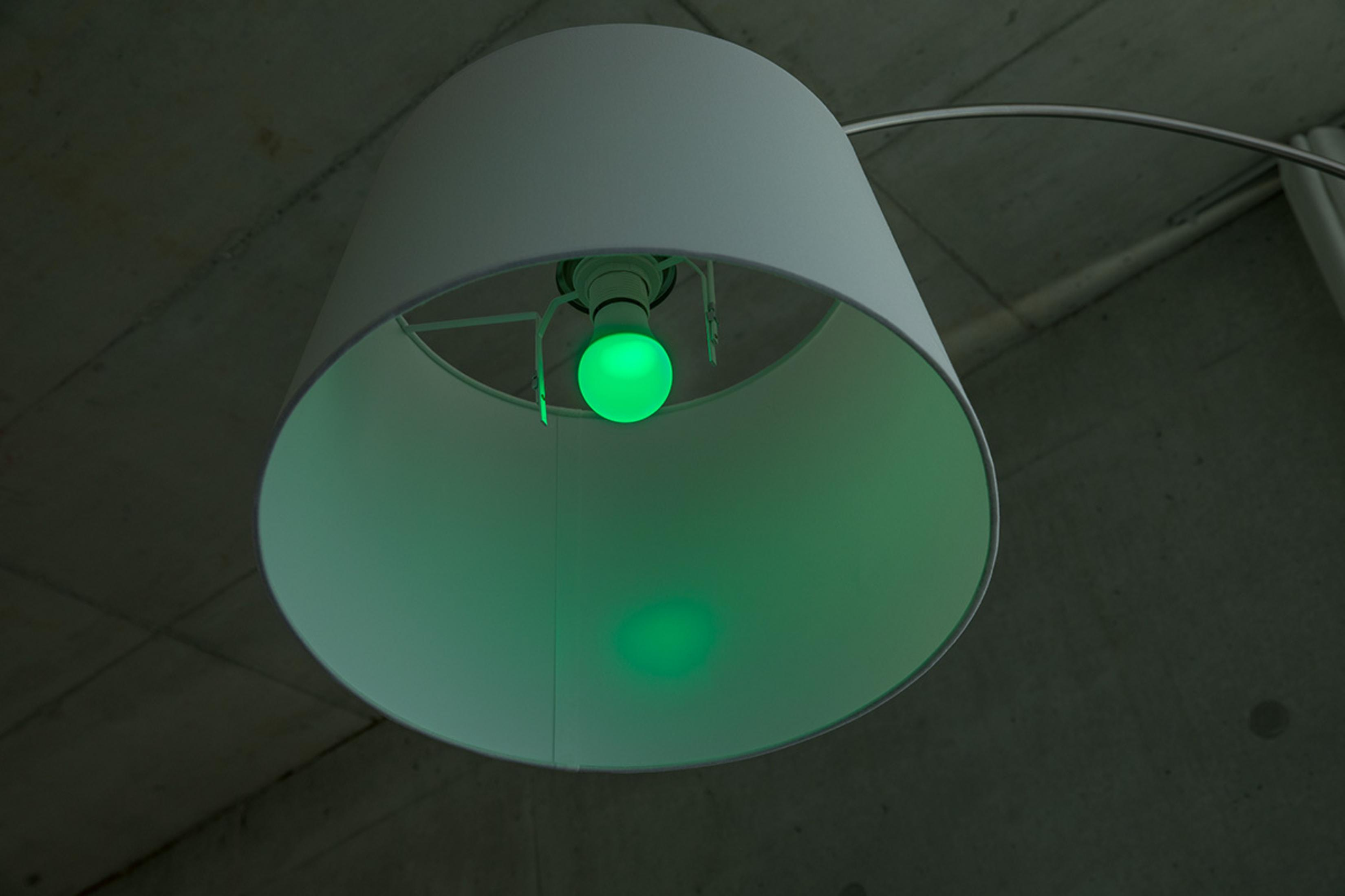 Lampadina verde brilla in paralume bianco, sotto soffitto in cemento.