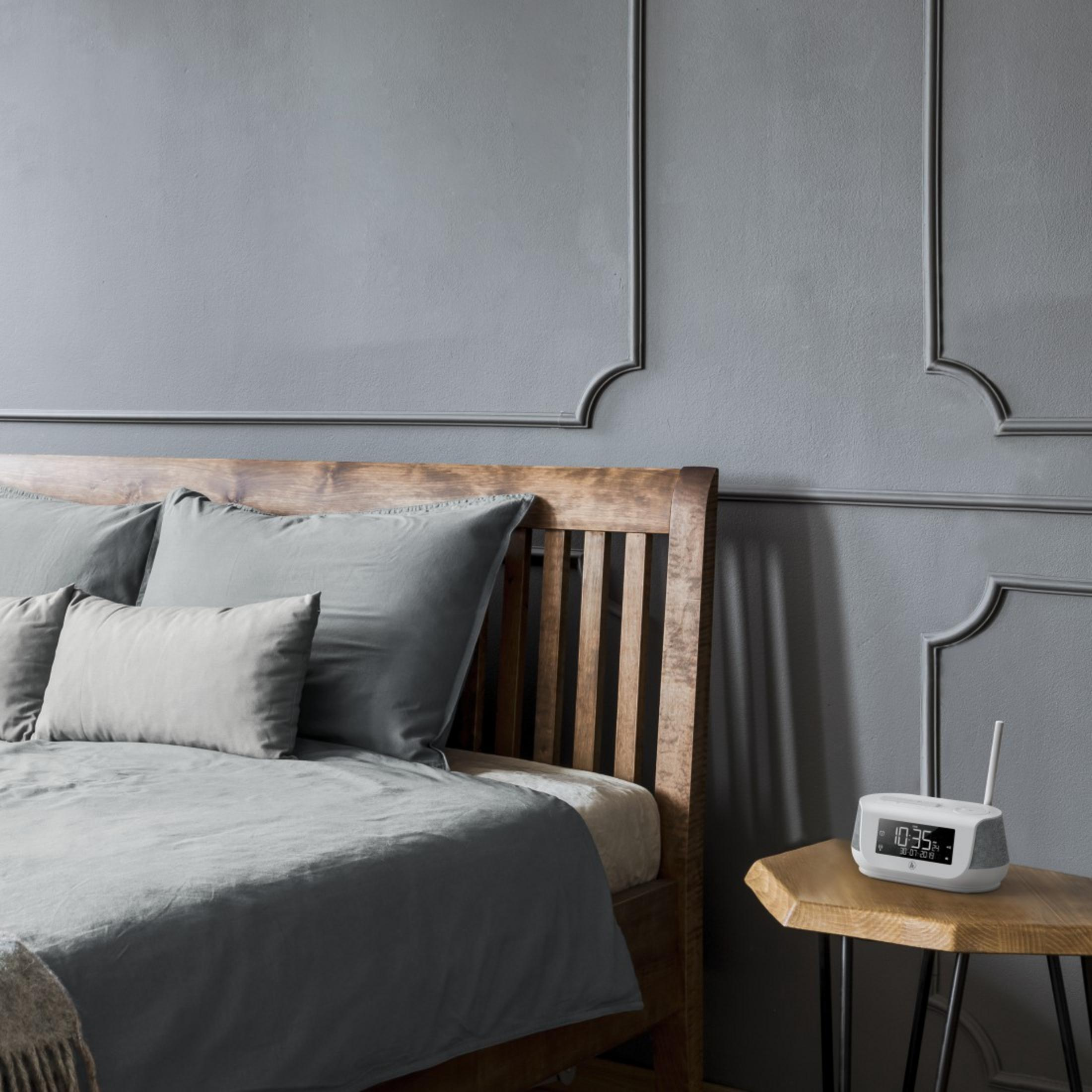 Scena sypialni z łóżkiem, poduszkami i radiem z budzikiem na stoliku. Szara ściana.