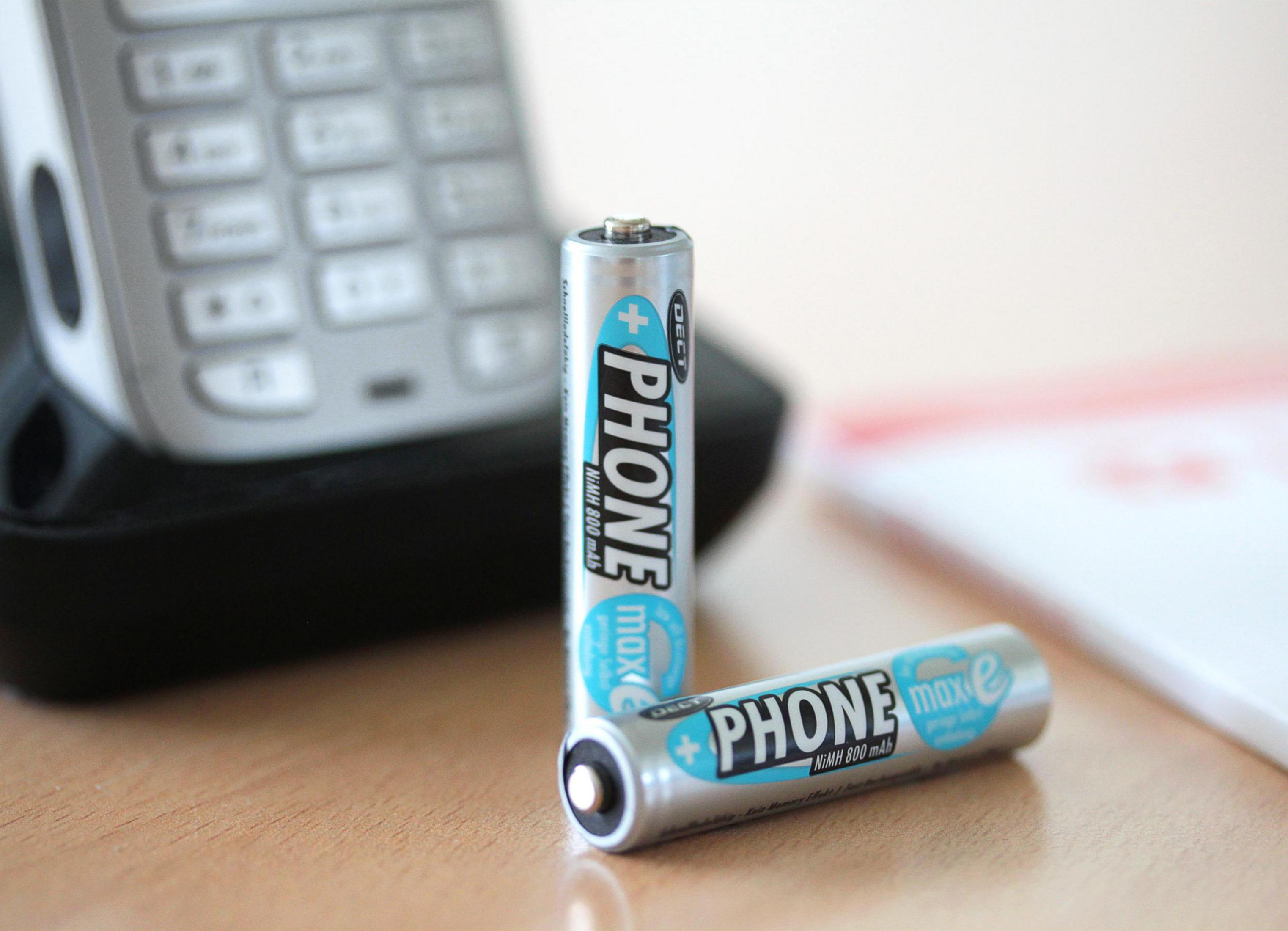 Zwei AA-Batterien neben einer Telefonbasis und einem Notizblock auf einem Holztisch.
