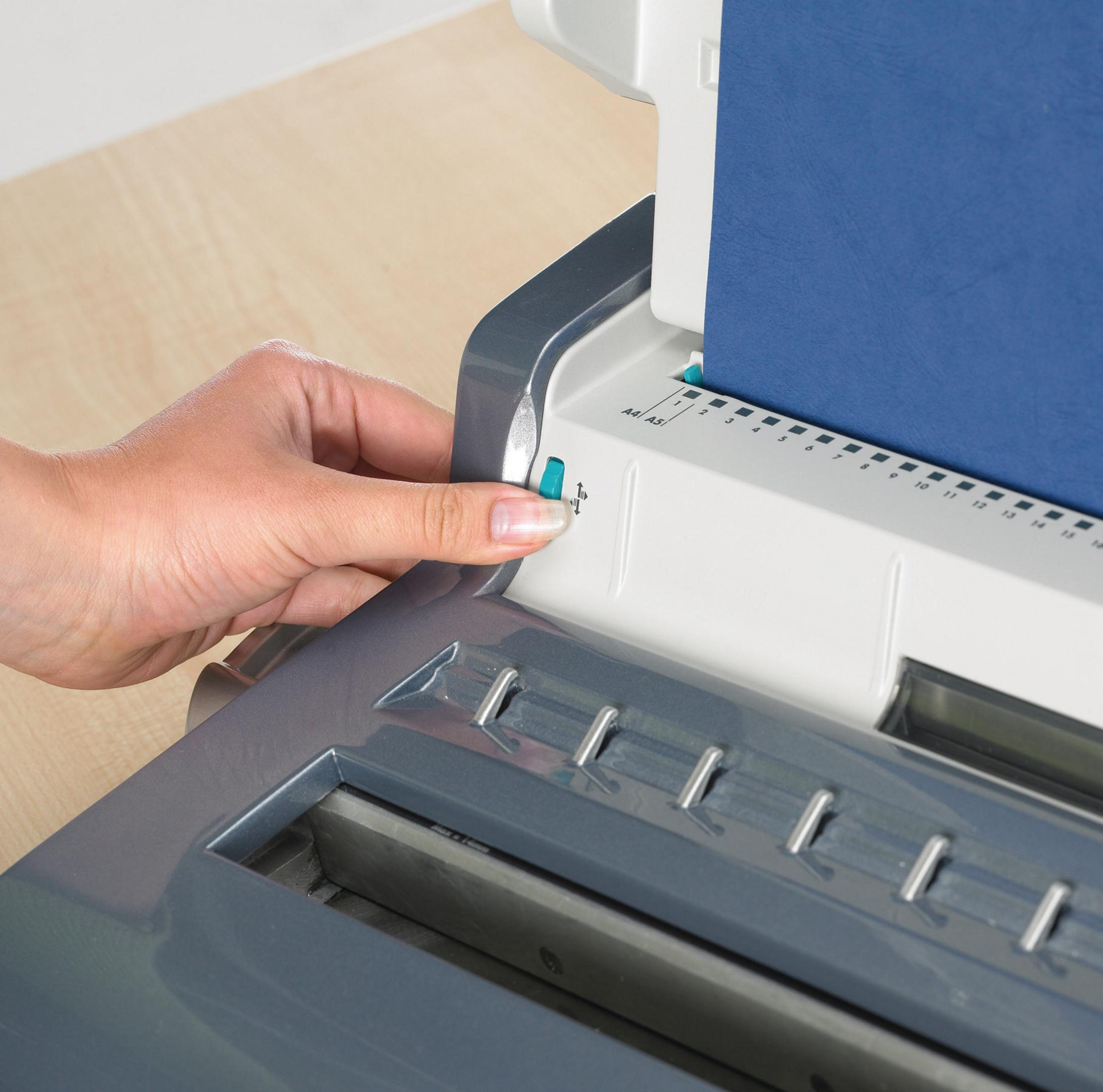 Eine Hand drückt einen grünen Knopf an einer grau-weißen Bindemaschine mit einem blauen Papierstapel.