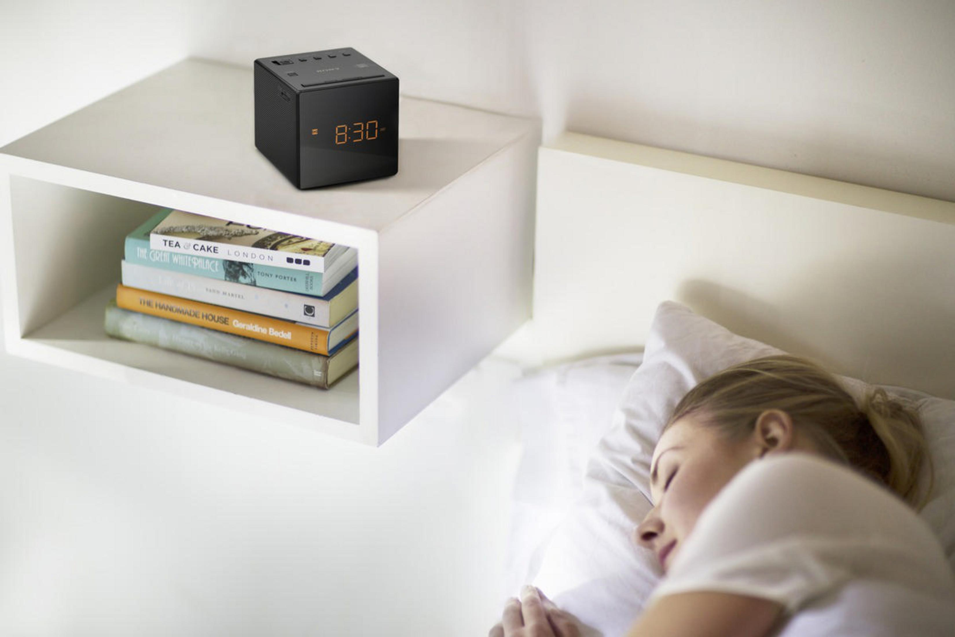 Ein schwarzer Wecker auf einem weißen Regal mit Büchern. Eine Frau schläft in einem Bett neben dem Regal.