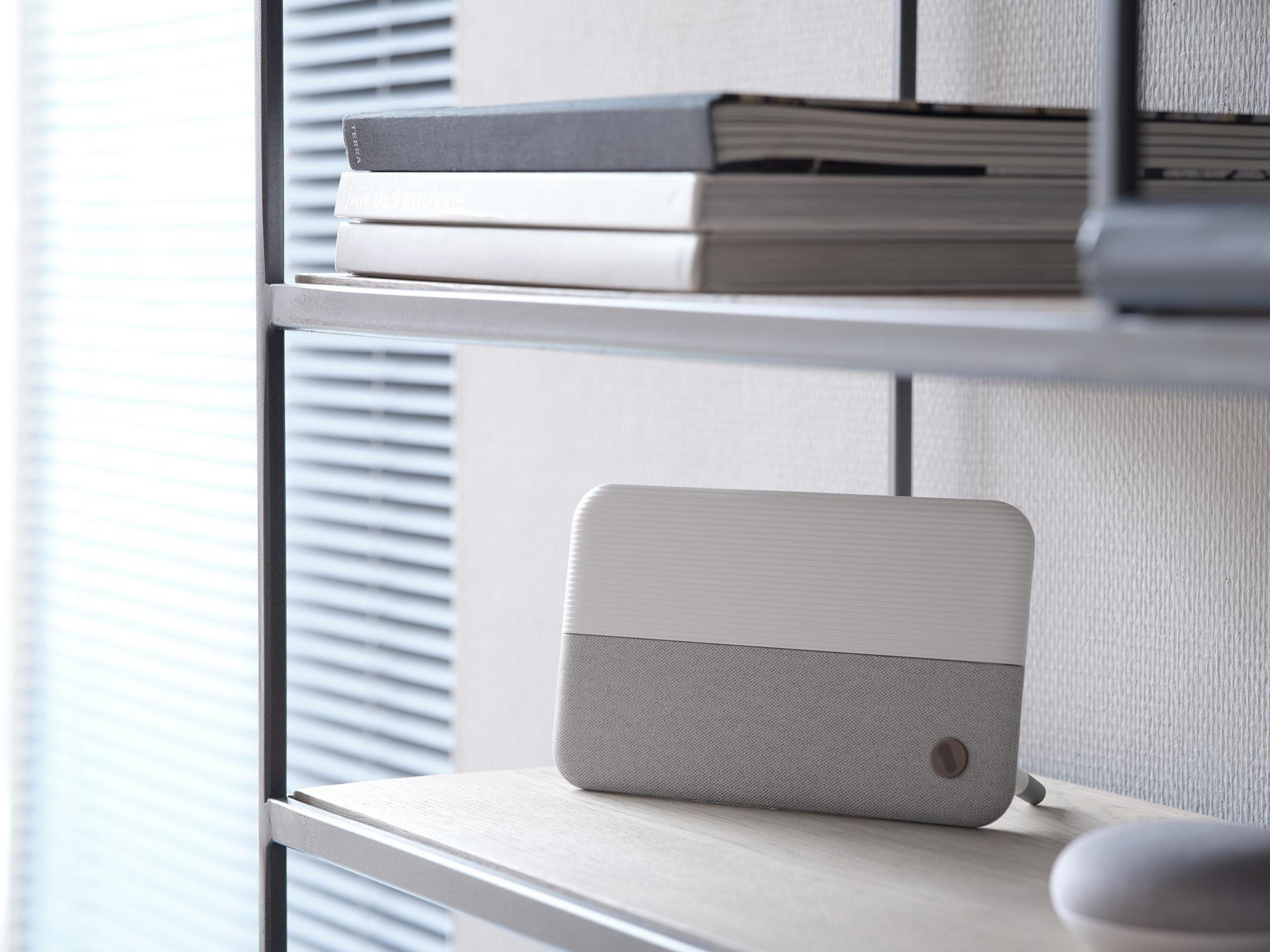 Biało-szary głośnik na półce z książkami i ścianą w tle.