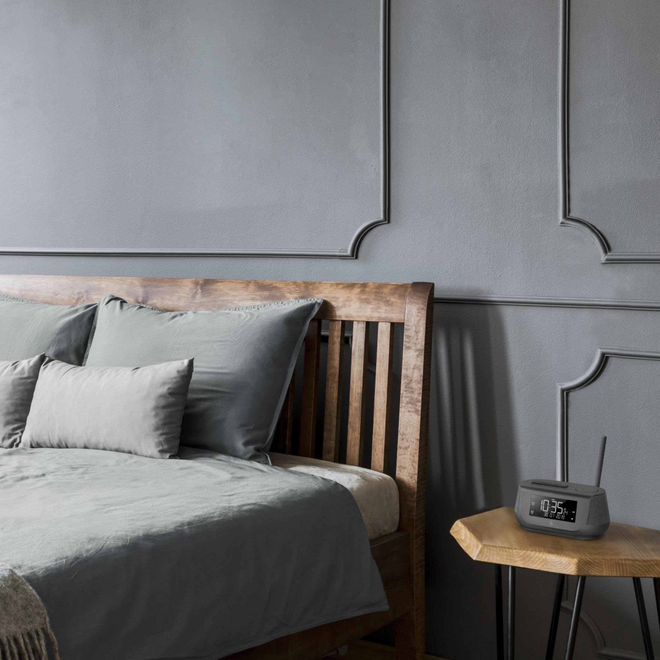 Scena sypialni: łóżko, poduszki, szara pościel, stolik nocny z zegarem.