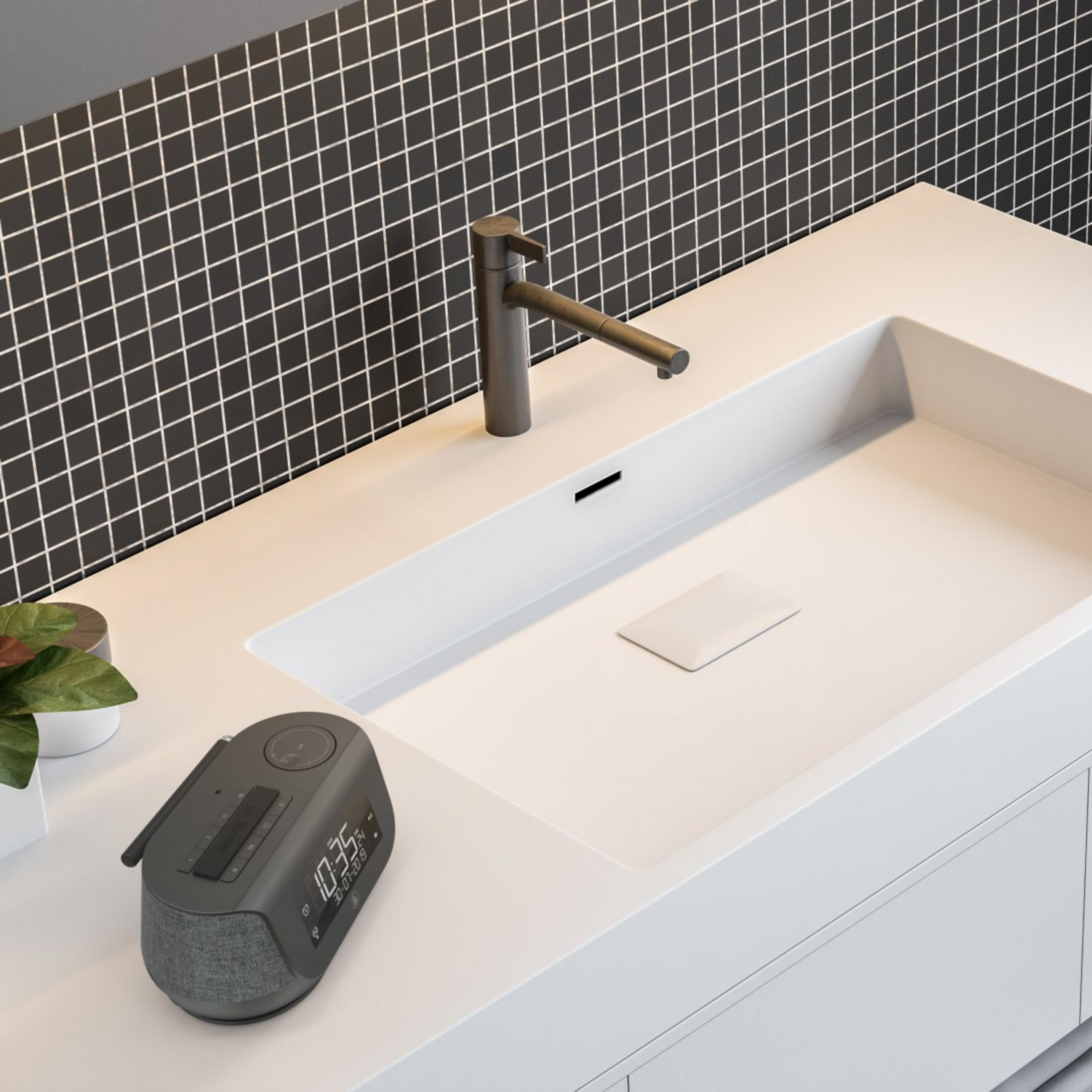 Wnętrze łazienki z białą umywalką, kranem i ciemnoszarą radiem z budzikiem. Czarne tło z płytek.