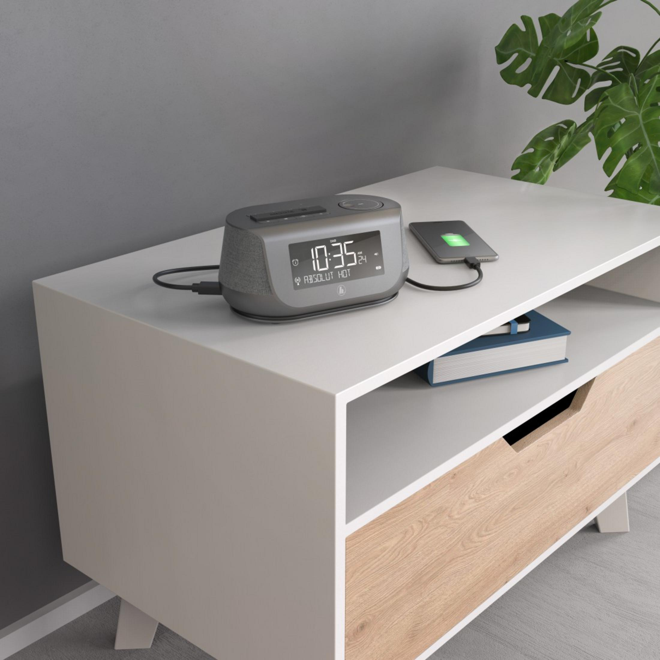 Szary budzik z telefonem ładującym się na wierzchu. Biała i drewniana szafka. Roślina w tle.