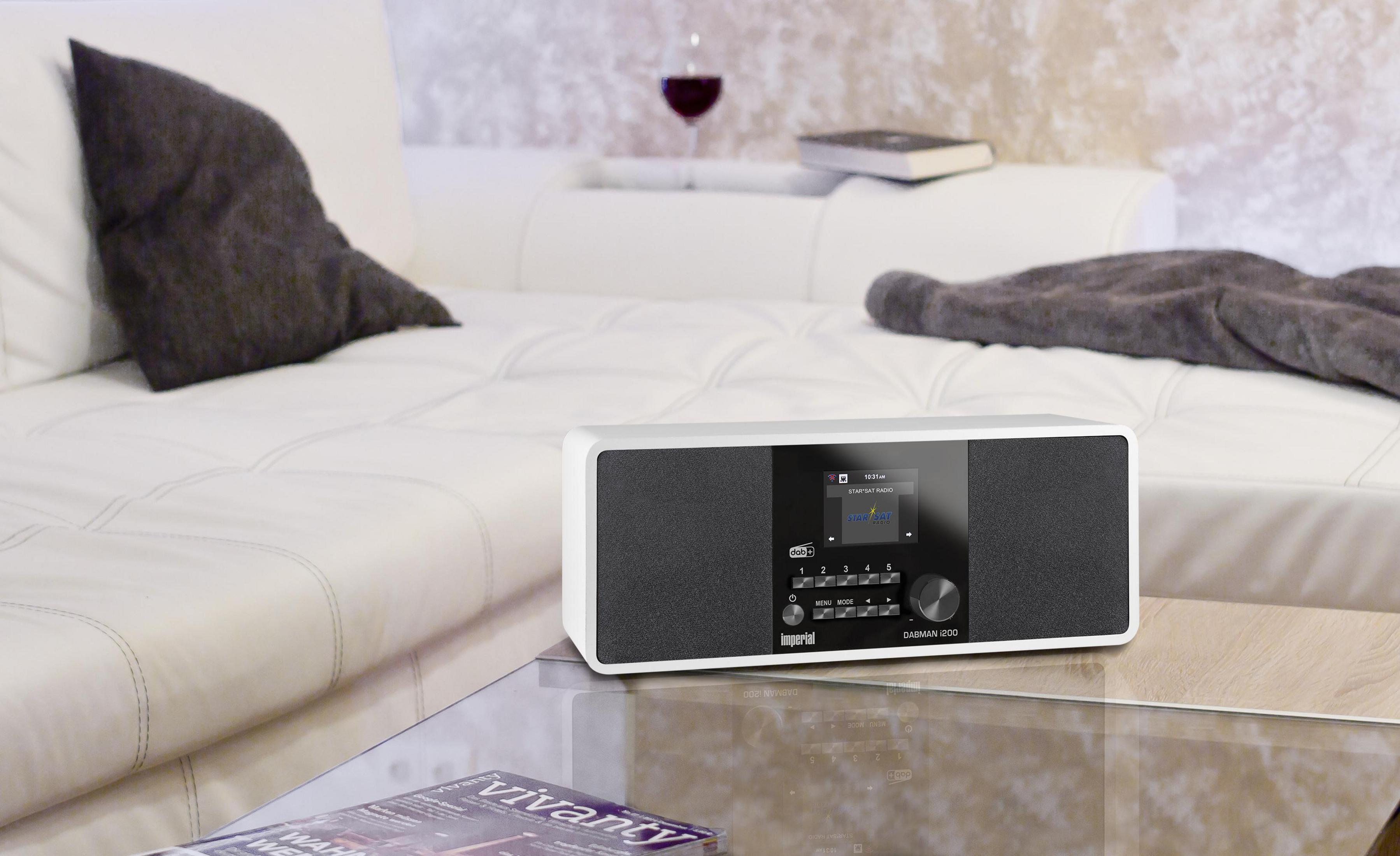 Biało-czarne radio z ekranem i przyciskami na szklanym stole. Kieliszek do wina i sofa w tle.