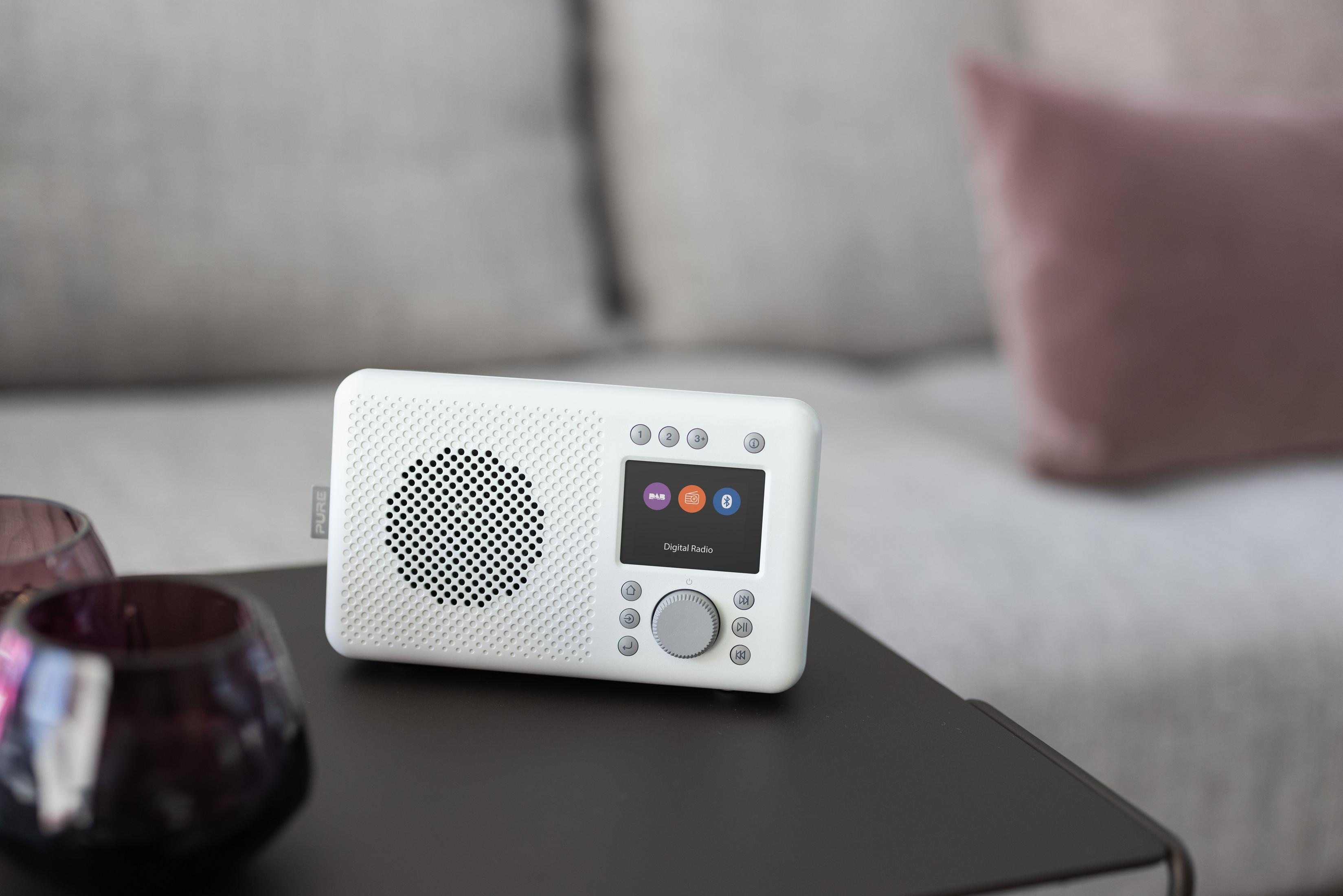 Białe radio cyfrowe na stole obok sofy, z ekranem i przyciskami.