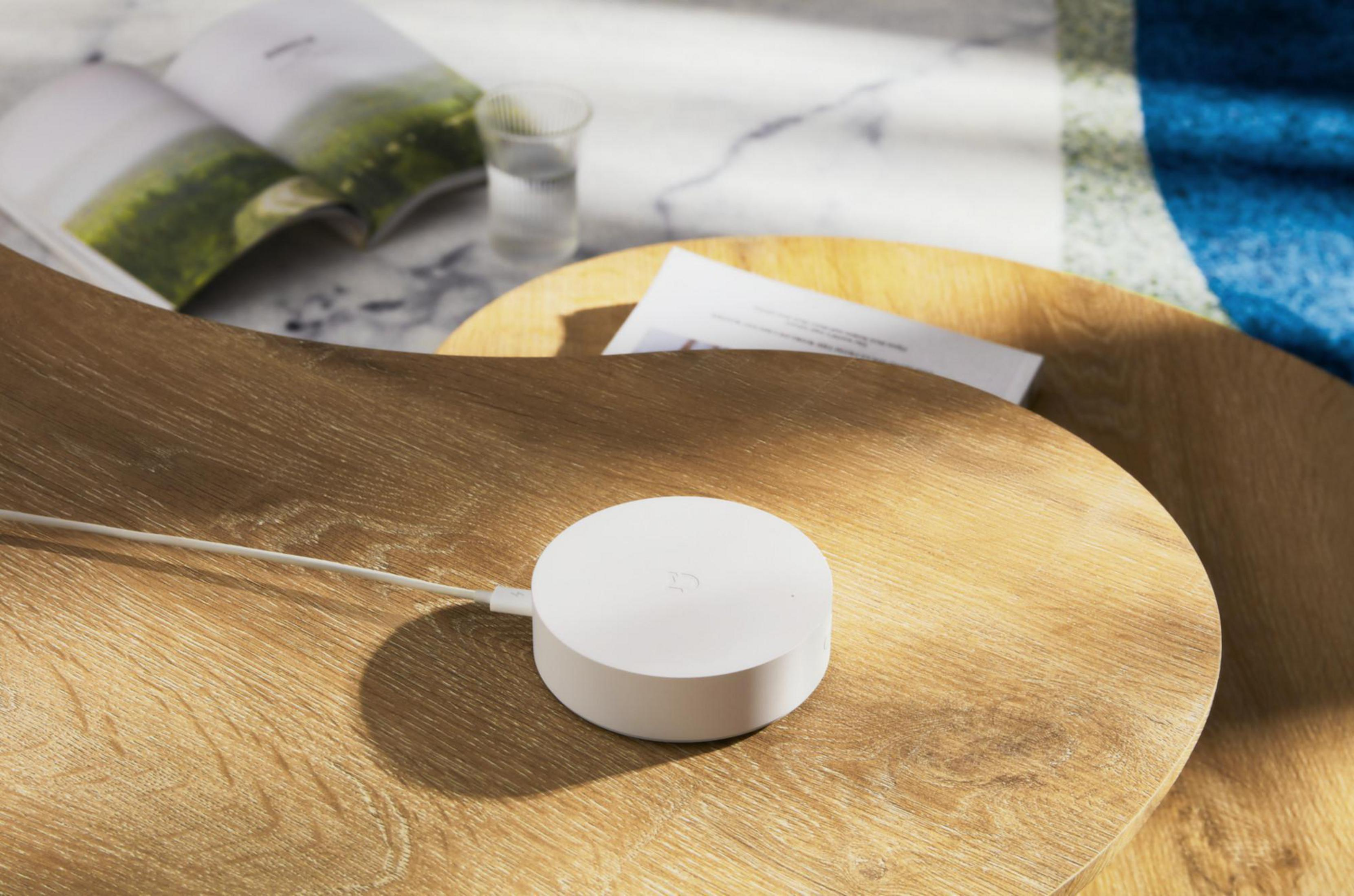 Un dispositivo smart home bianco su tavolo di legno, con bicchiere e libro aperto.