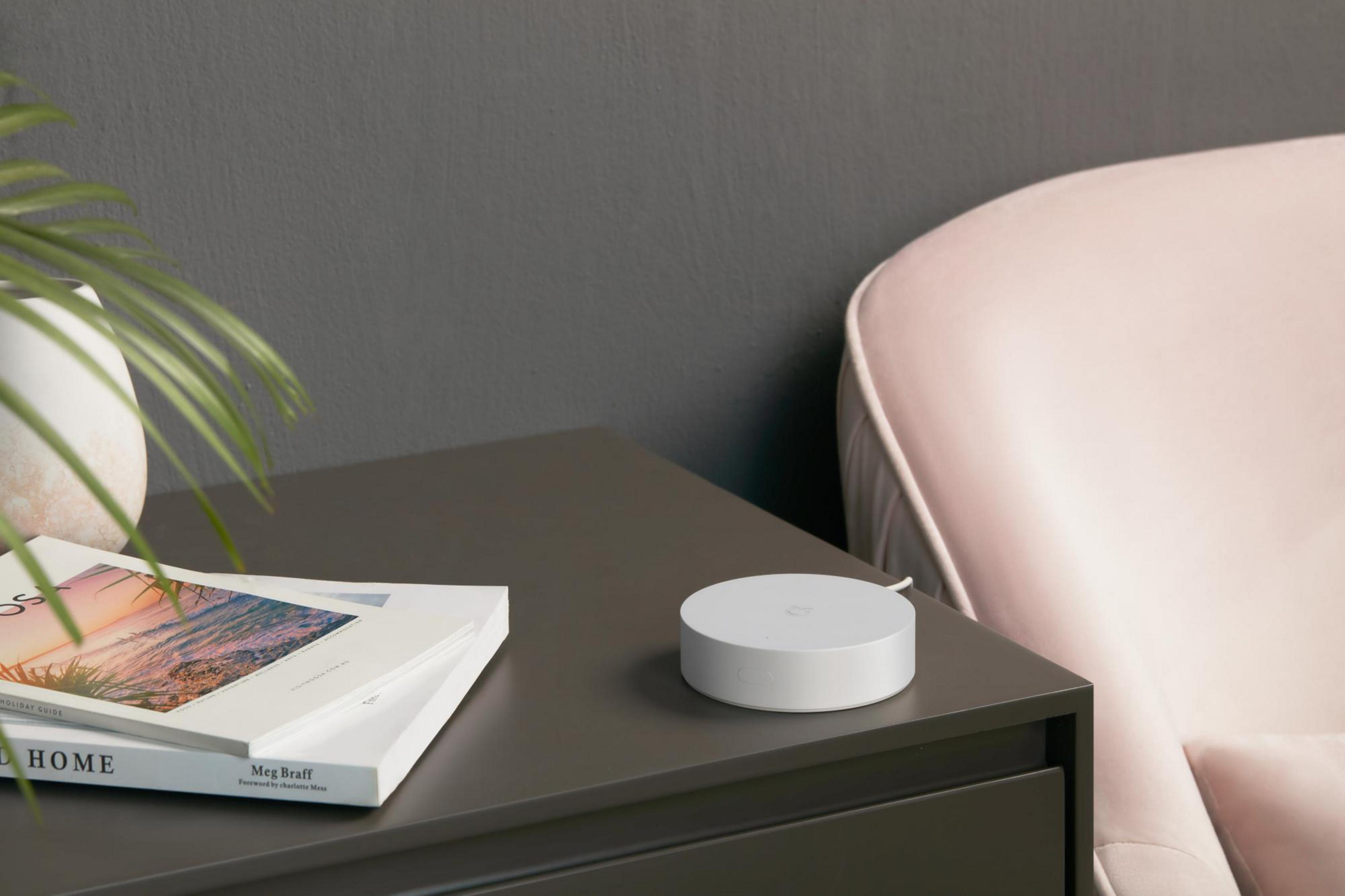 Router bianco su tavolo grigio scuro, rivista, pianta e sedia rosa sullo sfondo.