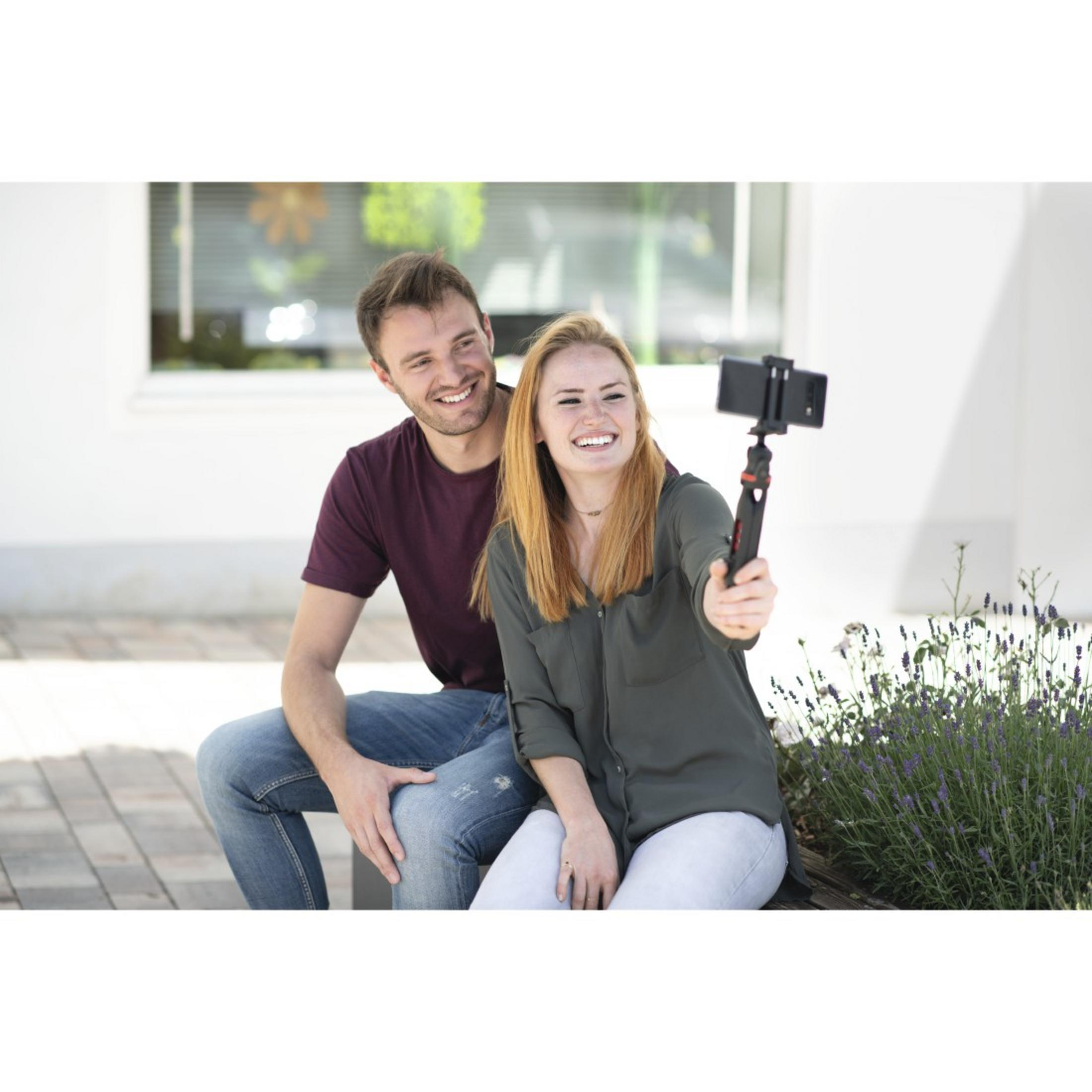 Una pareja haciéndose un selfie con un smartphone en un palo selfie, sonriendo al aire libre.