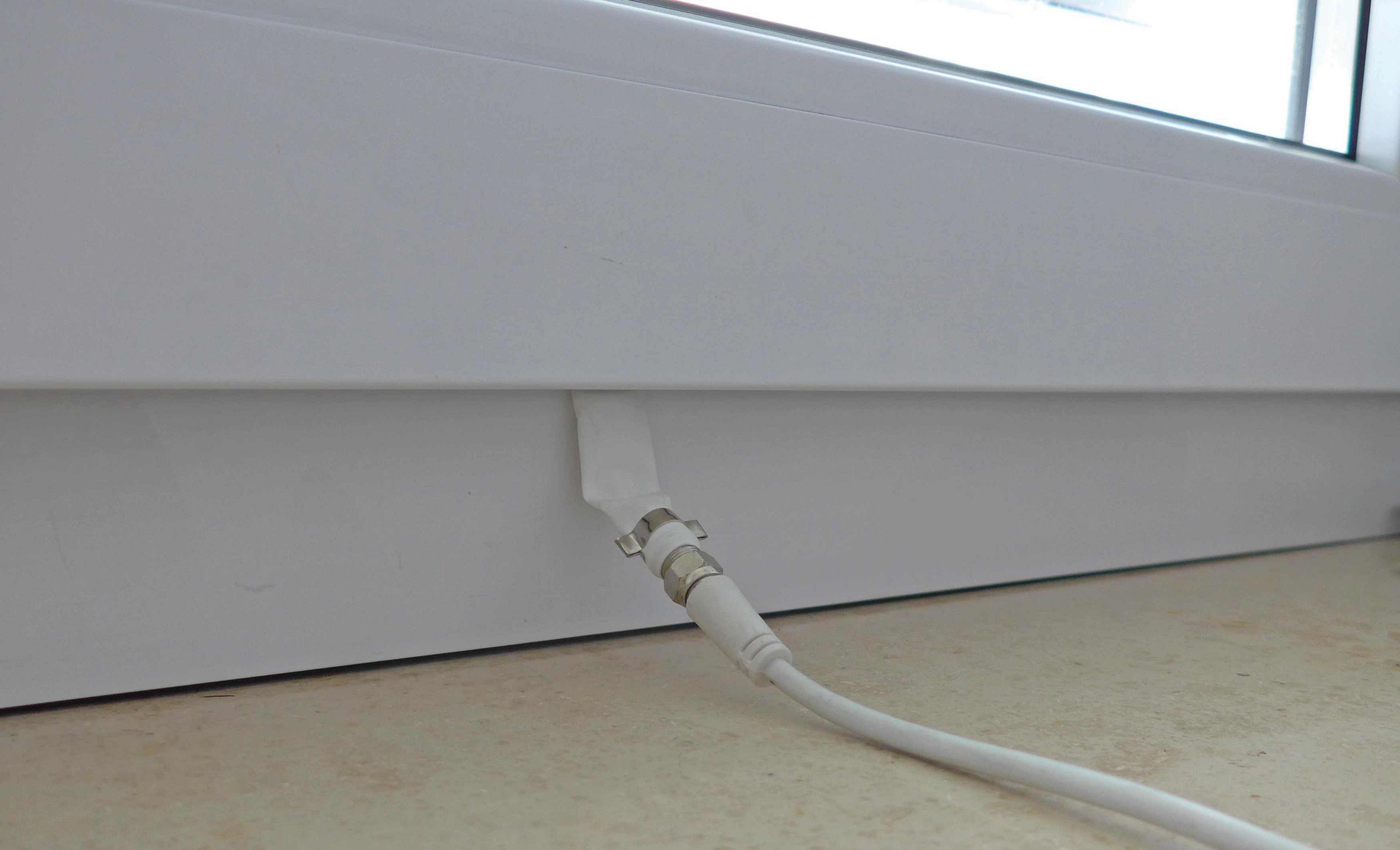 Ein weißes Kabel ist an einer weißen Wand befestigt. Das Kabel kommt von einer weißen Wand und ist in einem Fenster.