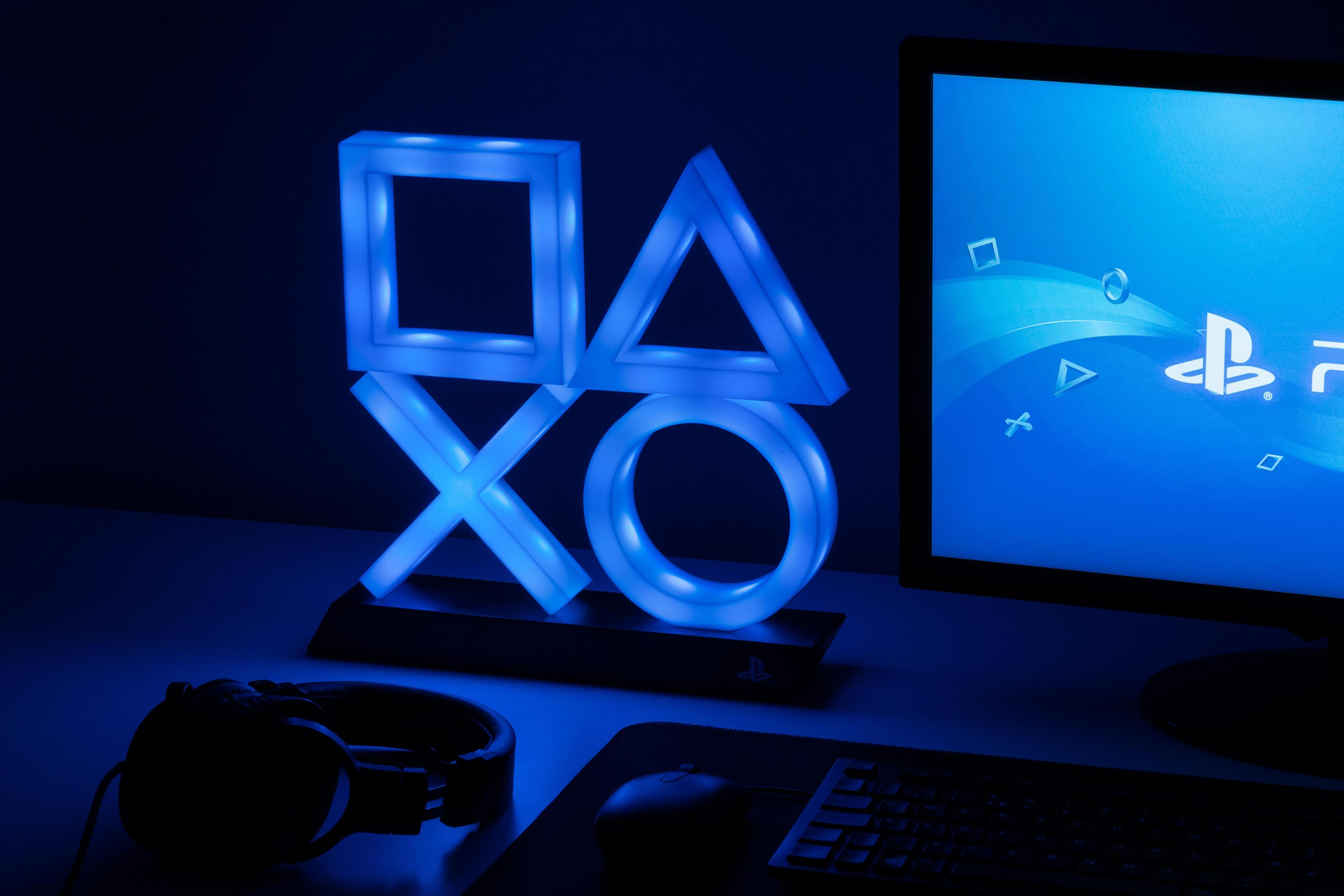 Leuchtendes Playstation-Symbol auf einem Schreibtisch. Ein Monitor und Kopfhörer sind ebenfalls sichtbar, alles in Blau.
