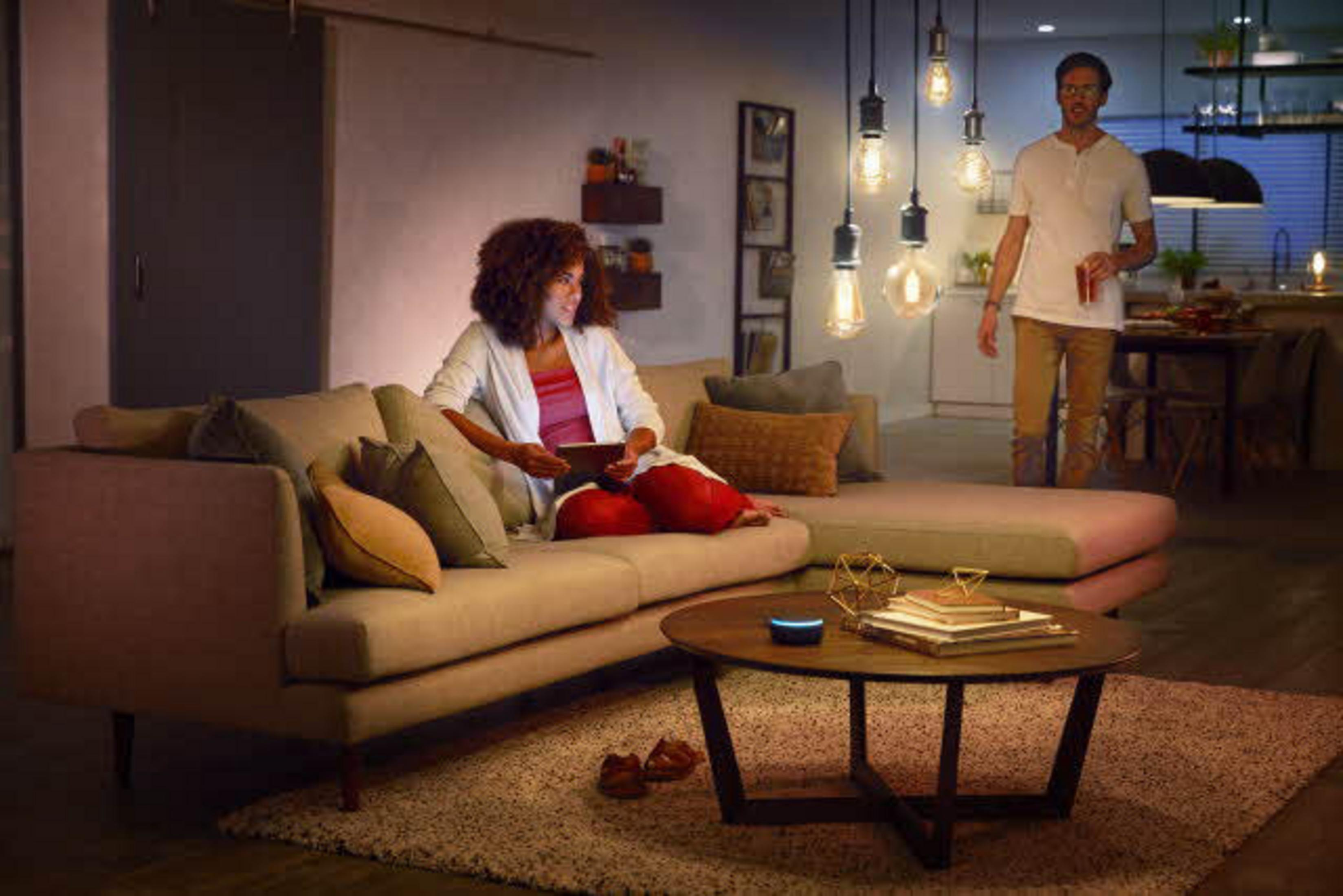 Scena w salonie z kobietą na sofie z tabletem, mężczyzną stojącym, oświetleniem otoczenia i okrągłym stołem.