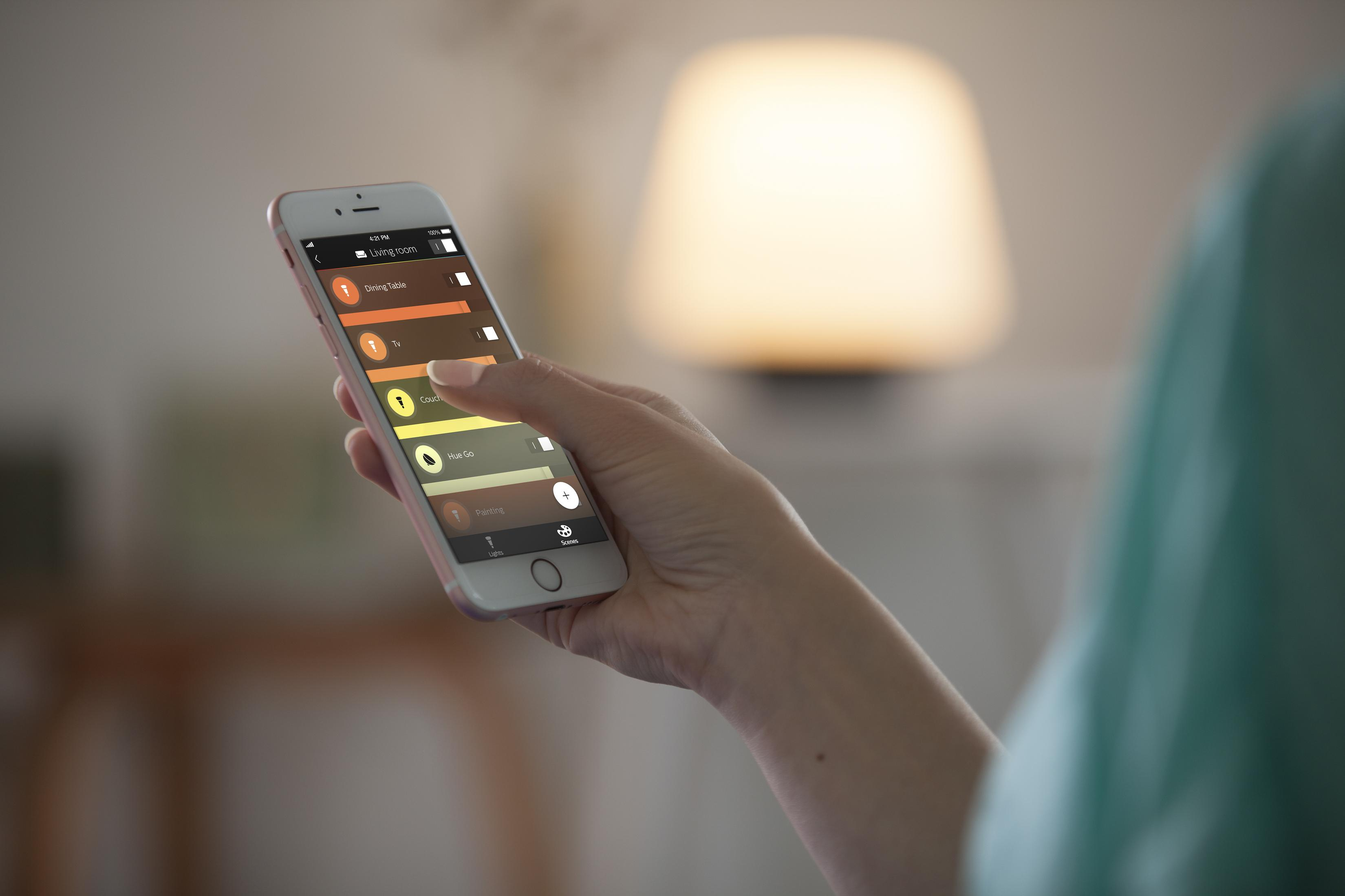 Die Hand einer Person, die ein Telefon hält, das eine Smart-Home-App anzeigt, in der Nähe einer Lampe.