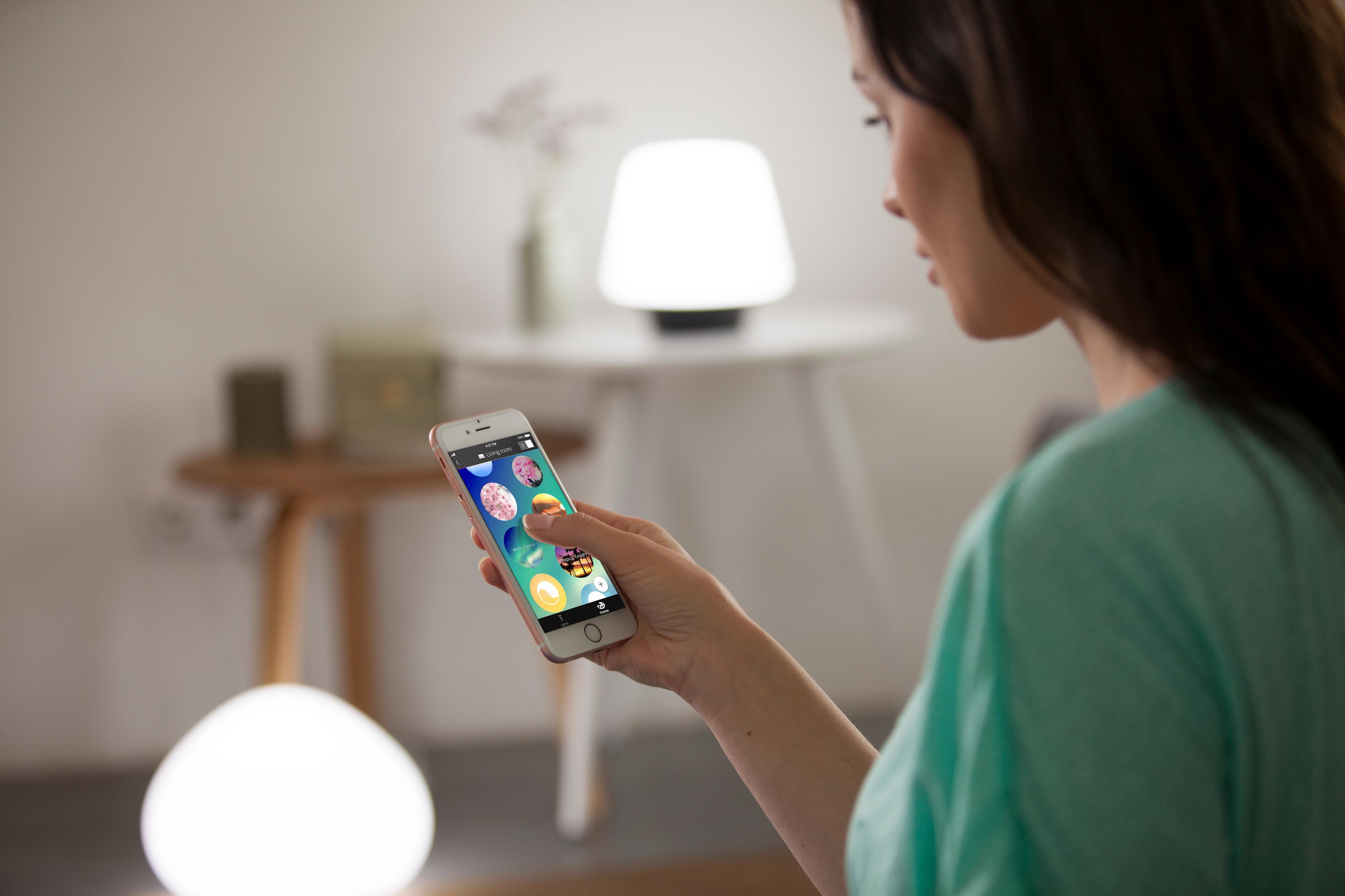 Eine Frau hält ein Telefon mit einer Smart-Lighting-App, eine Lampe steht auf dem Tisch.