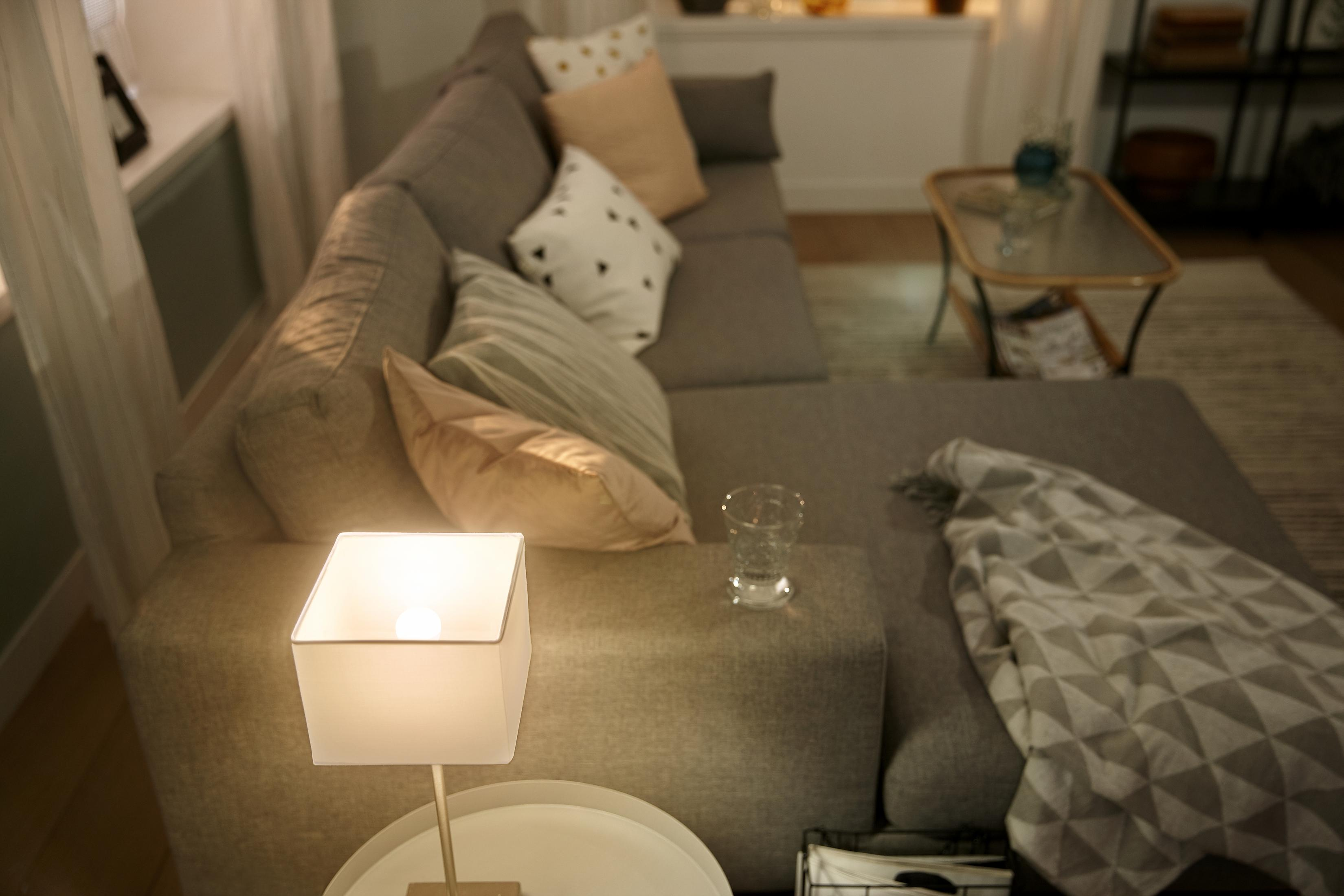 Szara sofa narożna z poduszkami i lampą na małym stoliku. W pobliżu stoi szklanka z płynem.