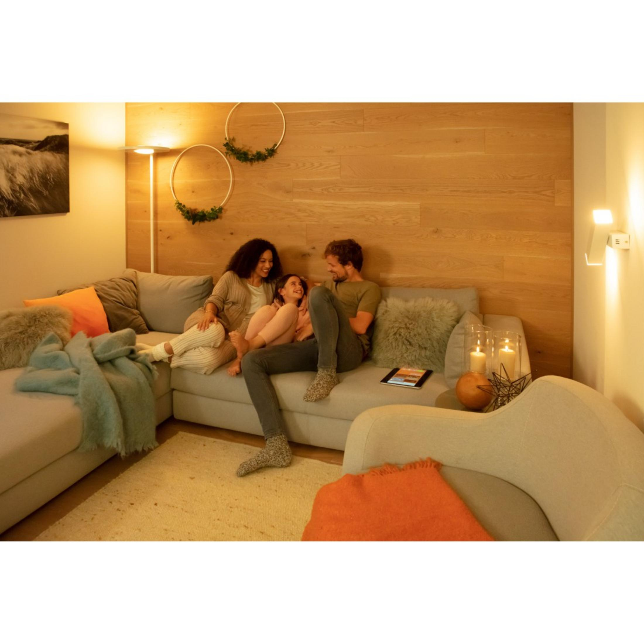 Drei Personen sitzen auf einem grauen Sofa. Es gibt eine Holzwand und eine Stehlampe. Gemütliches Wohnzimmer.
