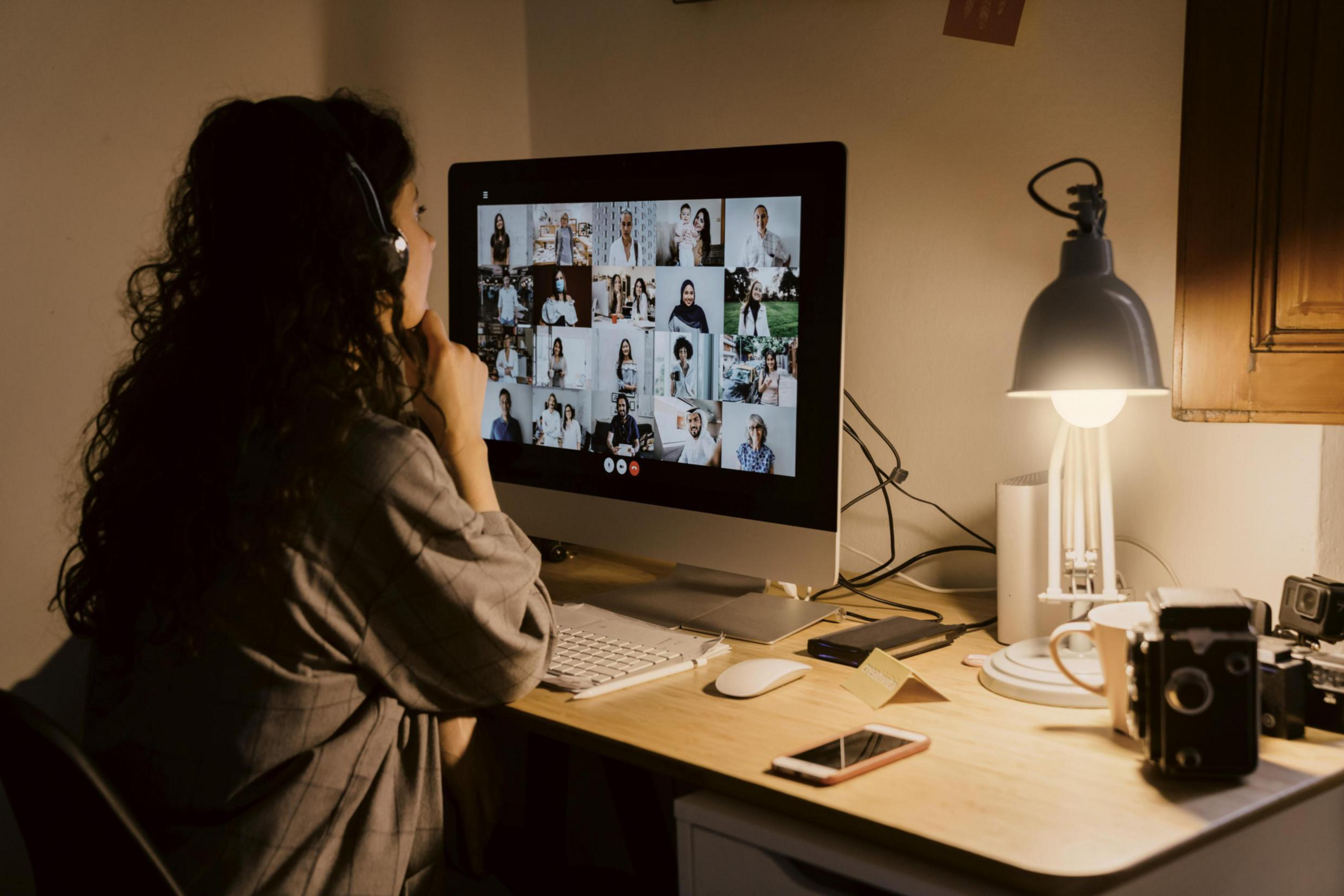 Donna in videochiamata. Uno schermo del computer mostra molti volti. Una scrivania con una lampada e un telefono.