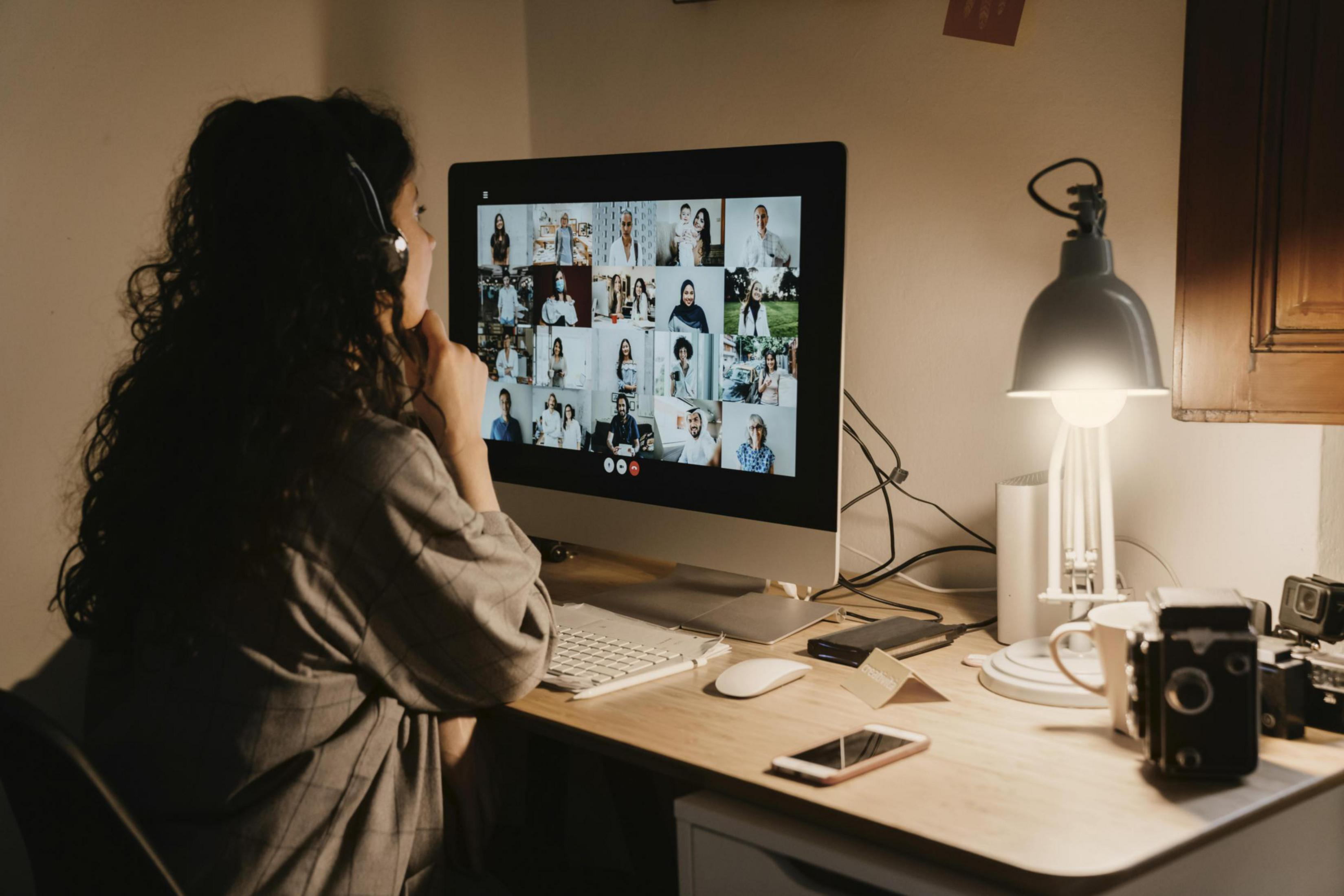 Una donna in videochiamata, che guarda lo schermo di un computer. Una lampada da scrivania e un telefono sono visibili.