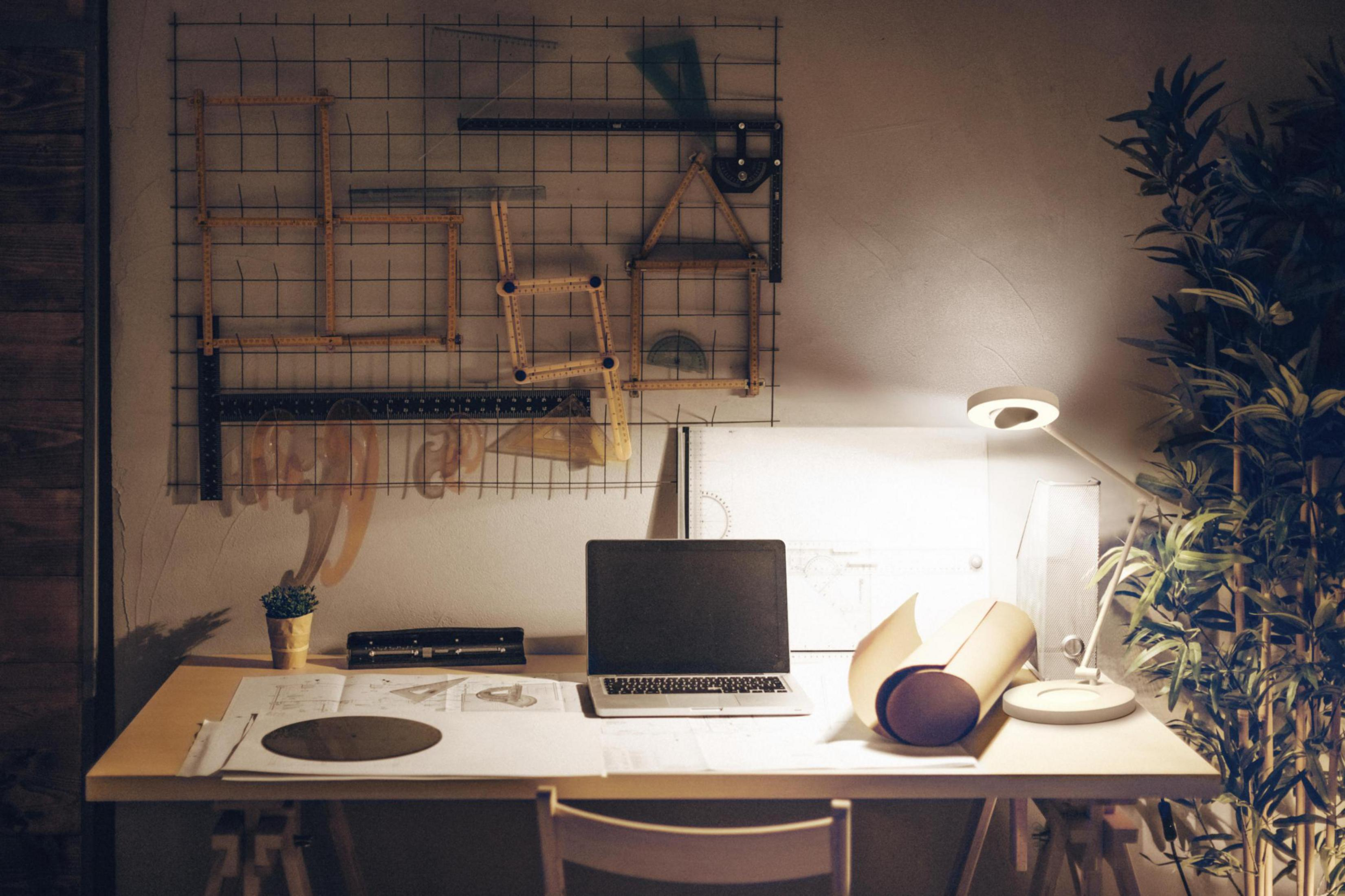 Schreibtisch mit Laptop, Bauplänen, Zeichenwerkzeugen, Lampe und Pflanzen.