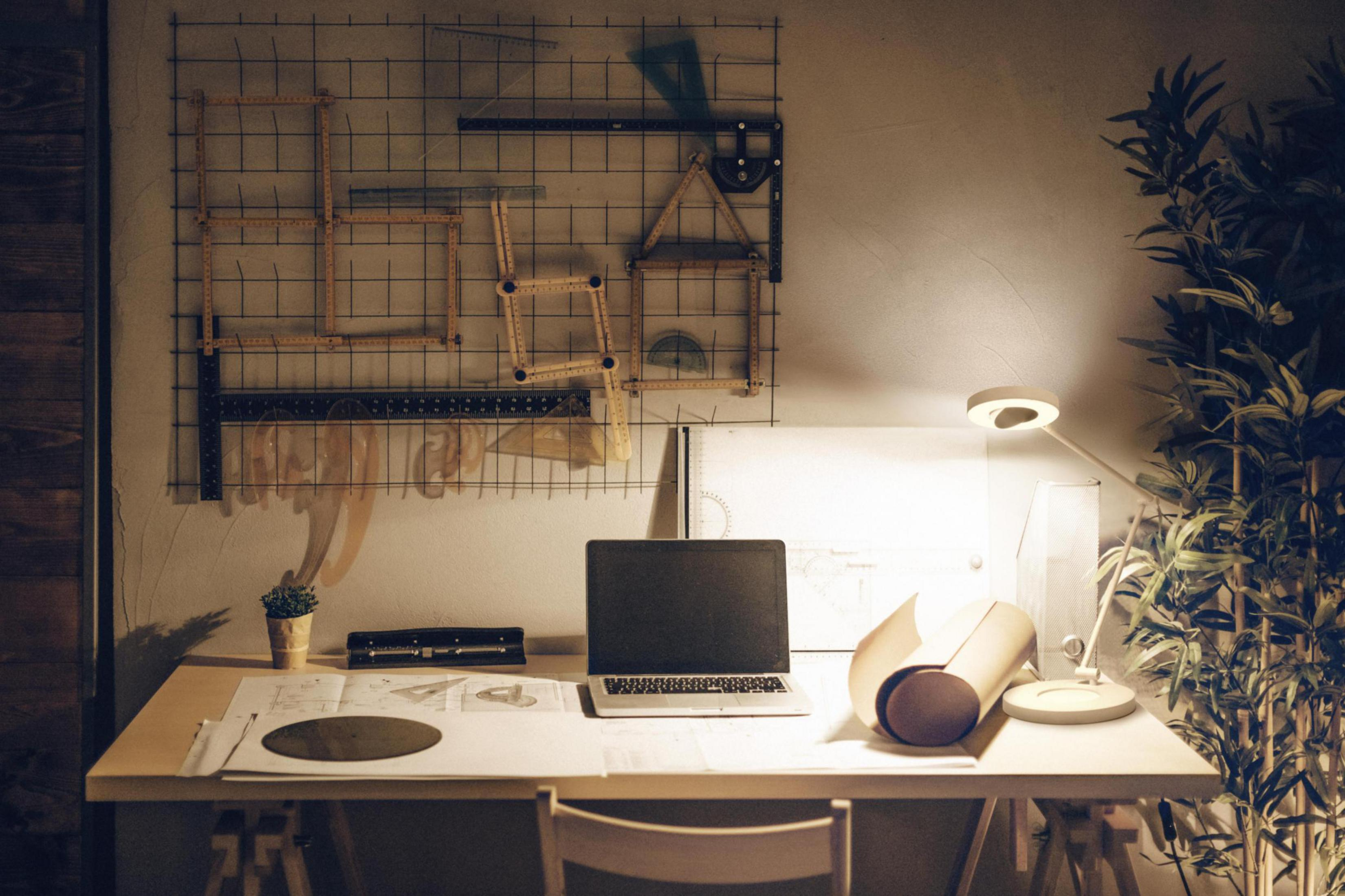 Ein Architektentisch mit Laptop, Blaupausen und Zeichenwerkzeugen, beleuchtet von einer Lampe.