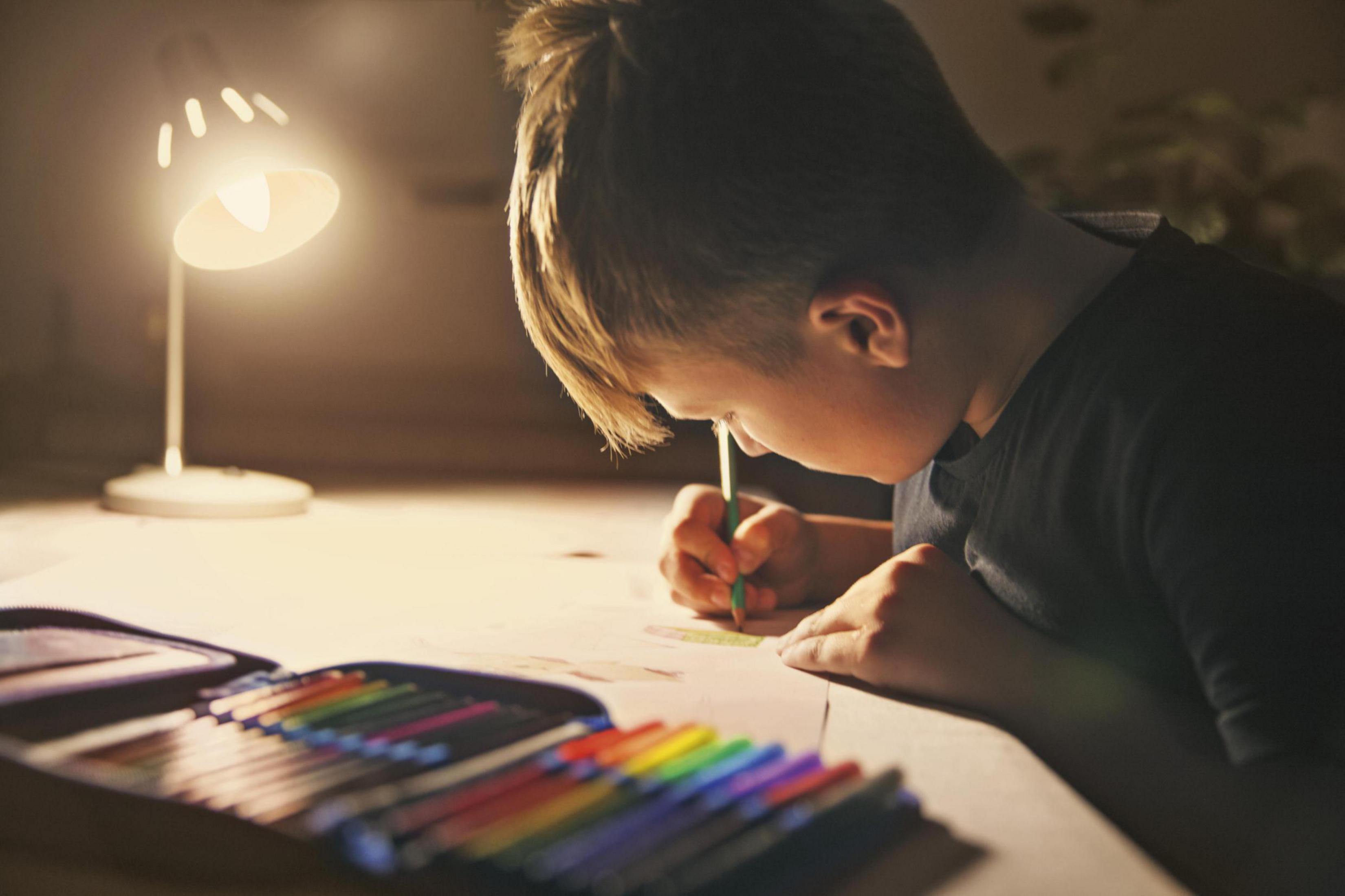 Chłopiec rysuje zieloną ołówkiem przy biurku, oświetlony lampą biurkową.