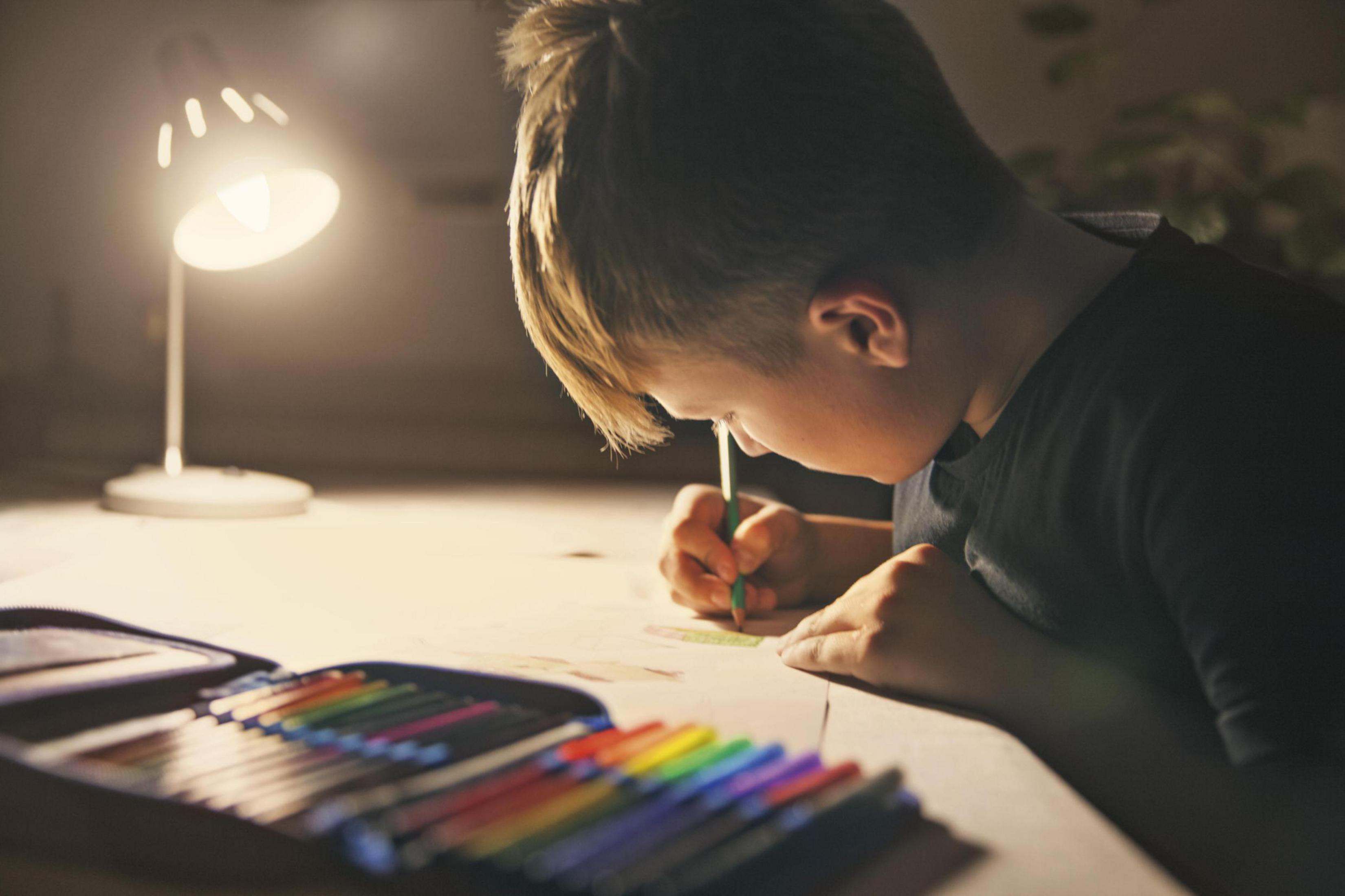 Chłopiec rysujący kredkami pod lampą biurkową.