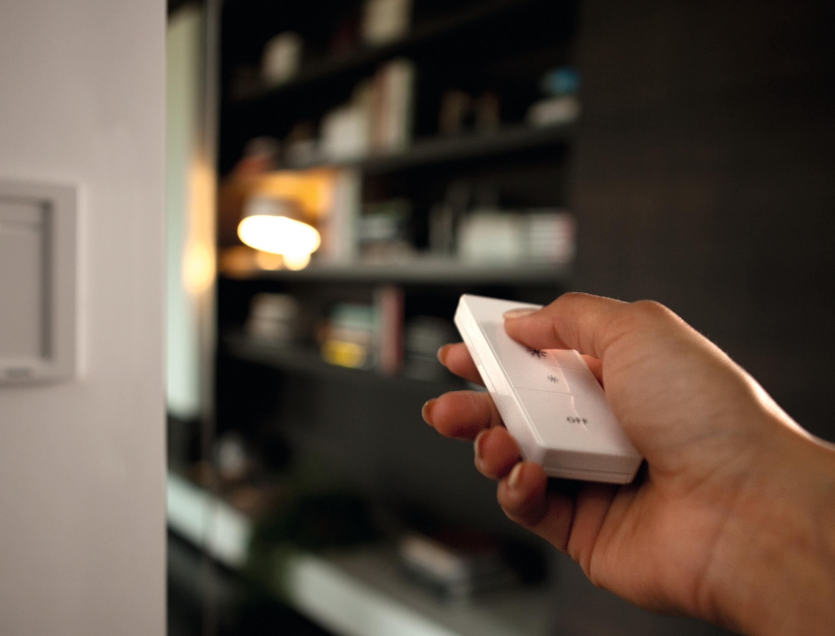 Hand hält eine weiße Fernbedienung. Der Hintergrund zeigt ein Bücherregal und eine Wand.