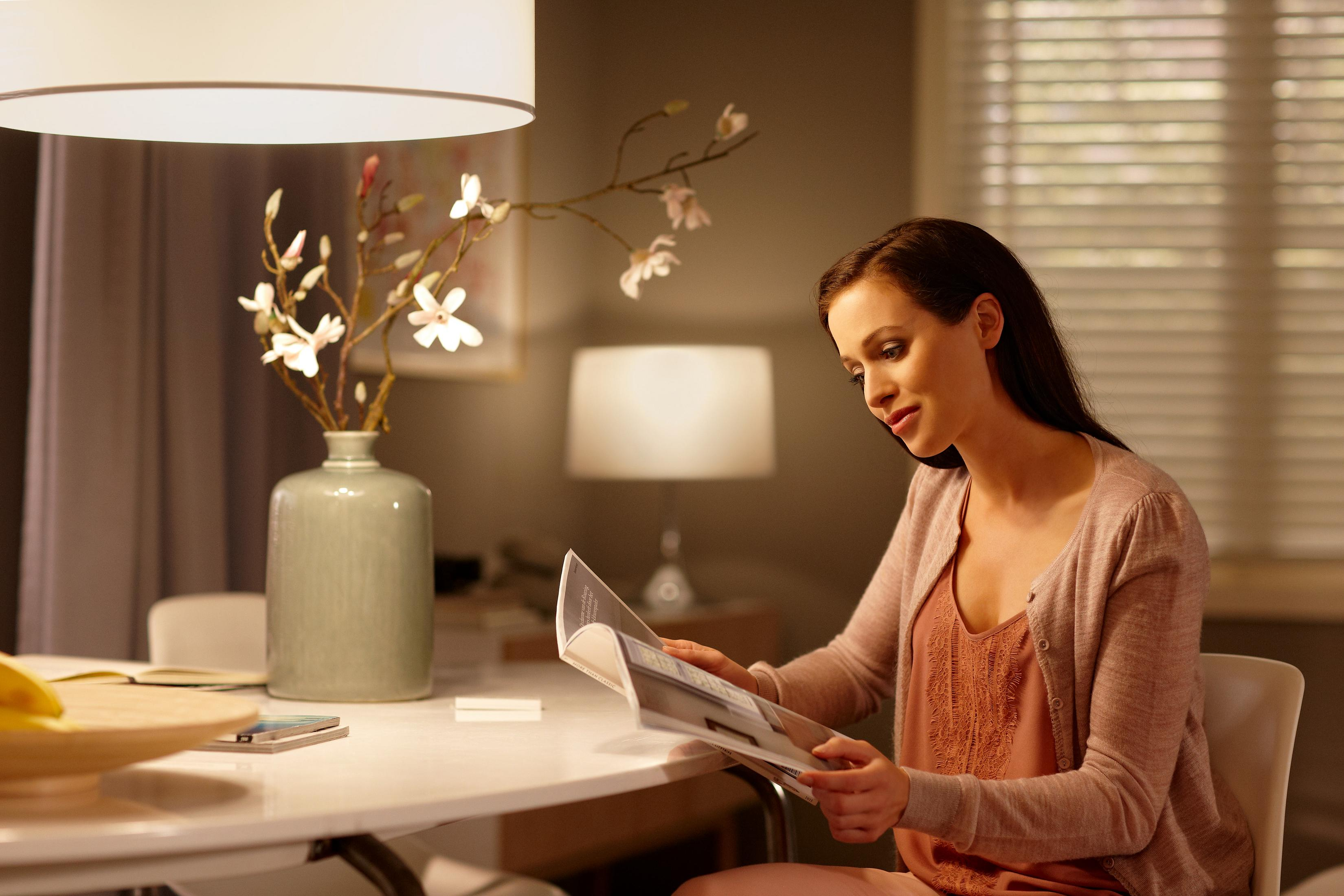 Frau liest eine Zeitschrift am Tisch, mit Lampe, Blumen und Fenster im Hintergrund.