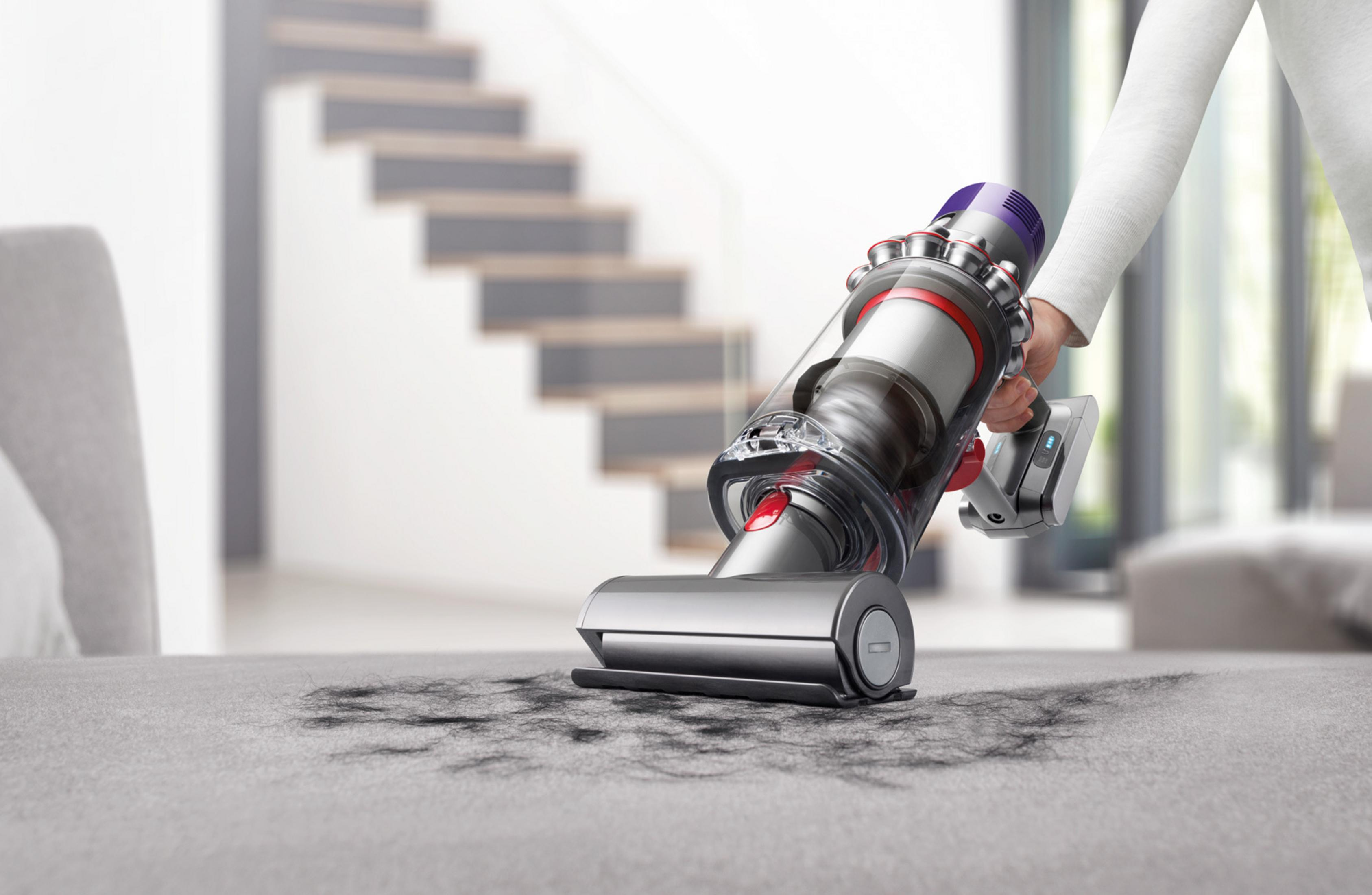 Eine Person saugt mit einem Dyson-Staubsauger ein graues Sofa ab und entfernt Haare und Schmutz.