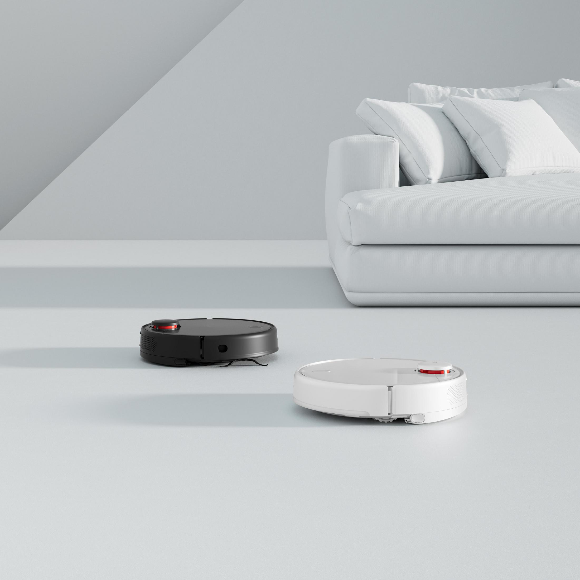 Açık bir zeminde iki robot süpürge. Bir siyah, bir beyaz, beyaz bir kanepenin yanında. Arka plan beyaz.