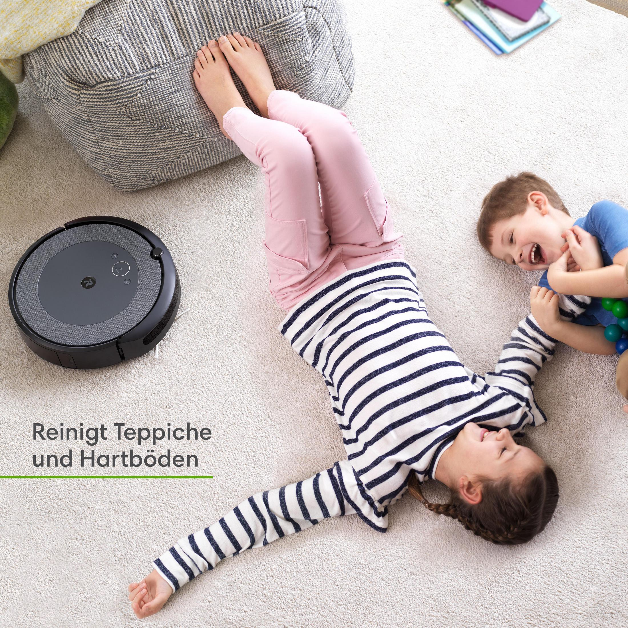 Zwei Kinder spielen mit einem Roboter-Staubsauger auf einem Teppichboden. Der Hintergrund hat ein graues Aufbewahrungsmöbel.