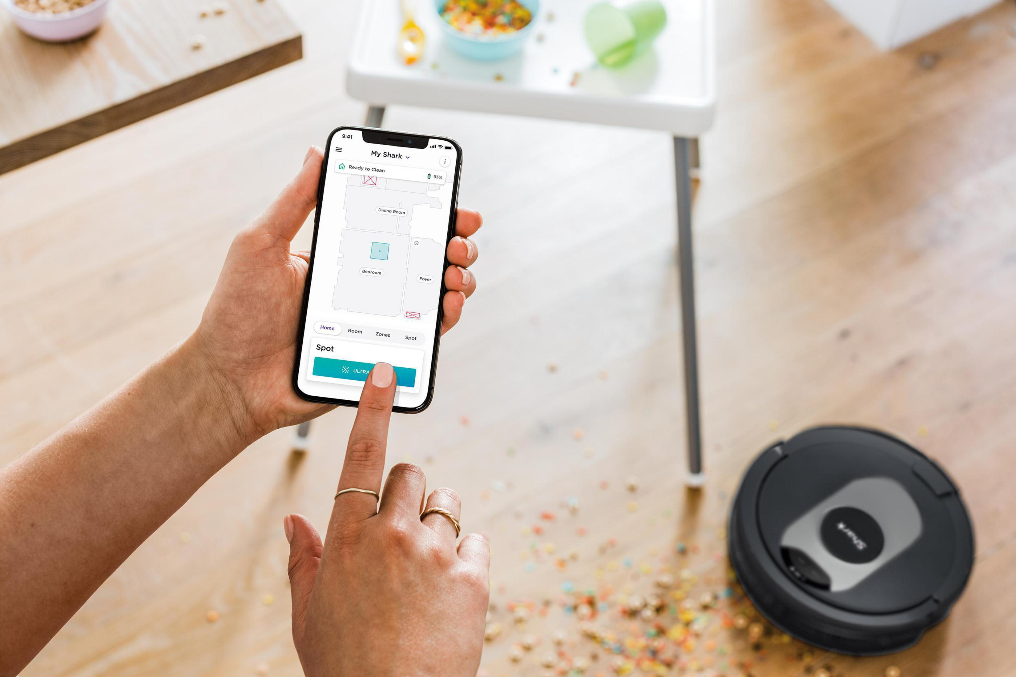 Ręce osoby używającej smartfona do sterowania robotem sprzątającym. Odkurzacz jest na podłodze.