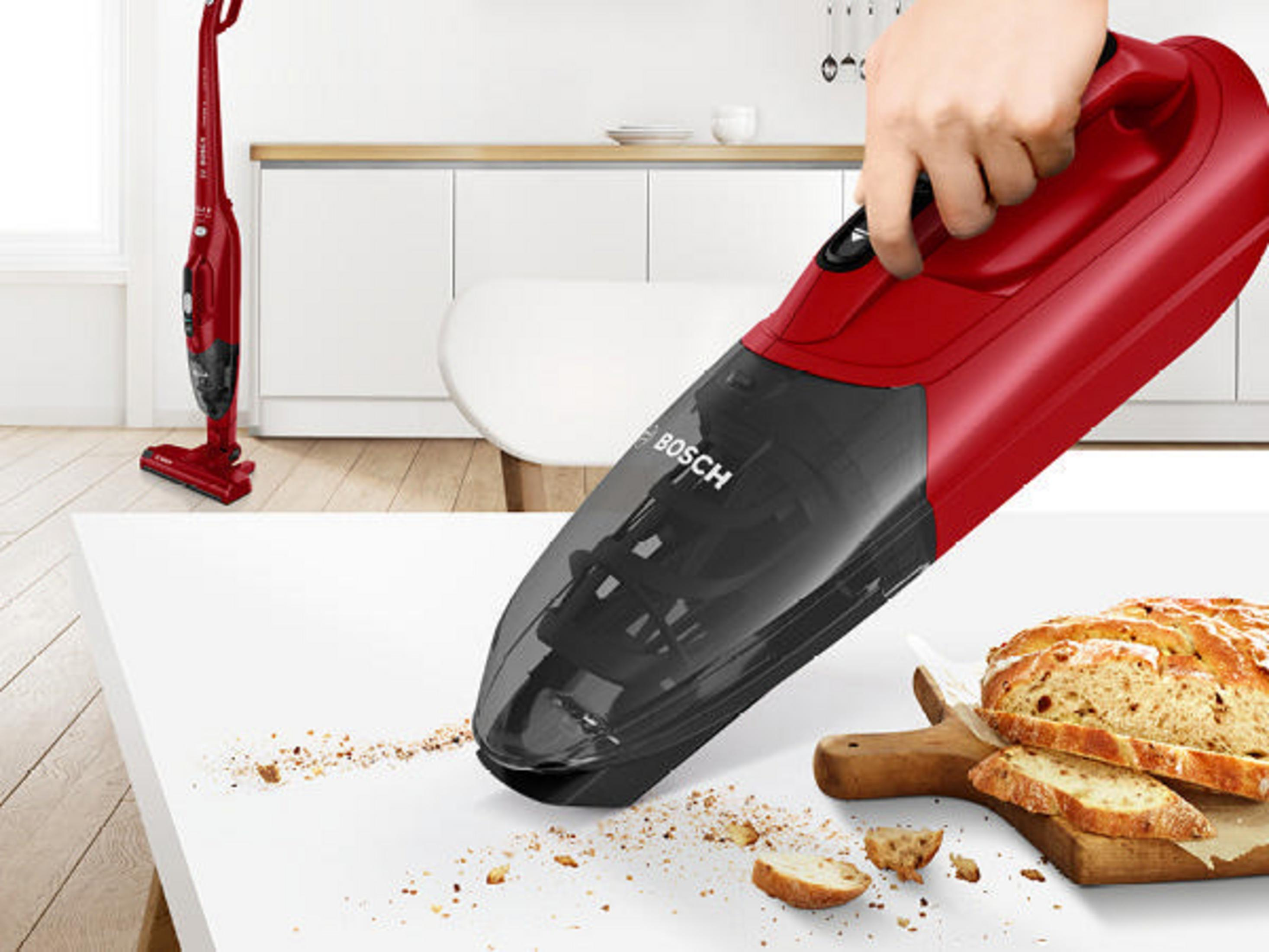 Czerwony odkurzacz ręczny czyszczący okruchy na białym stole obok chleba i deski.