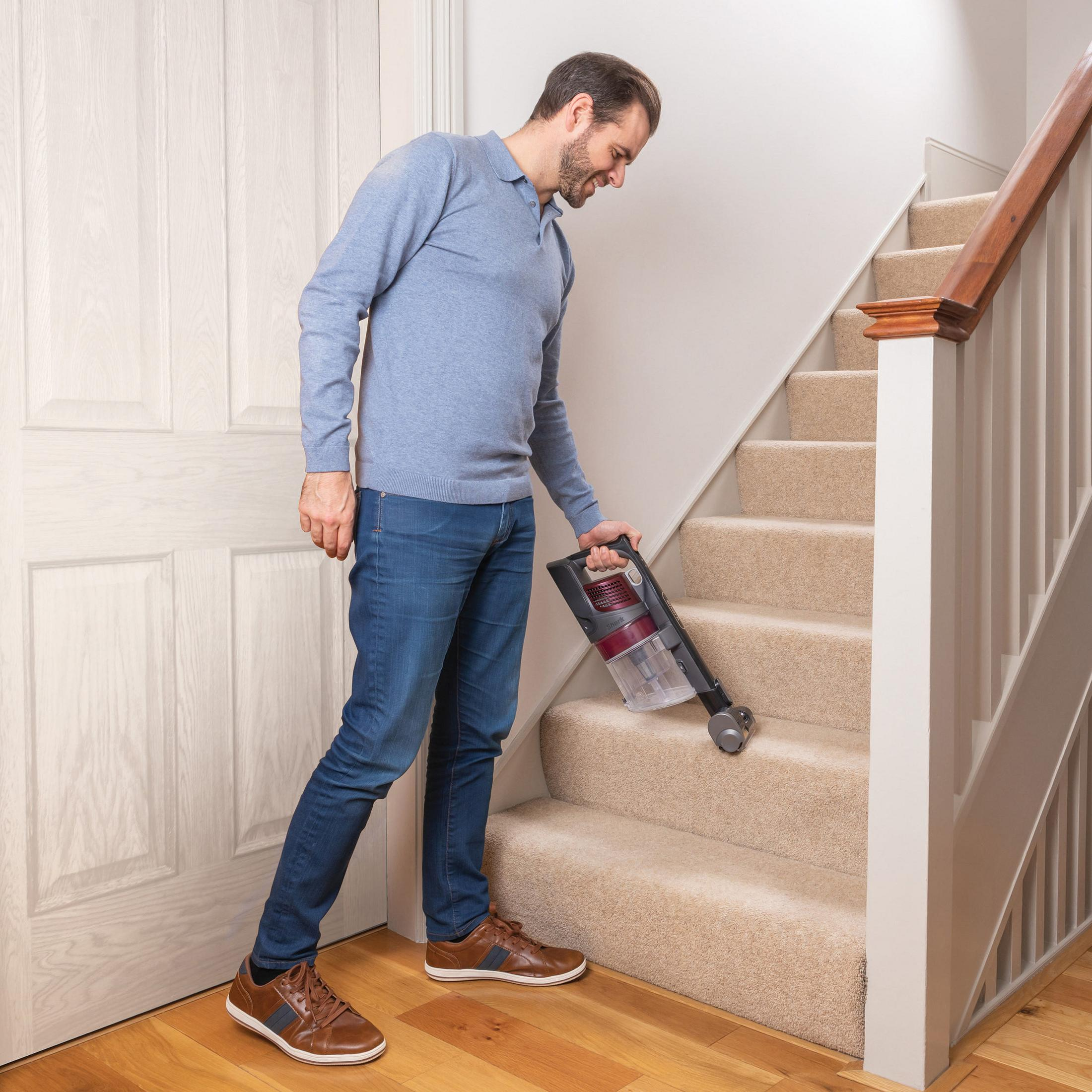 Un hombre aspira las escaleras alfombradas con una aspiradora de mano, sujetándola con la mano derecha.