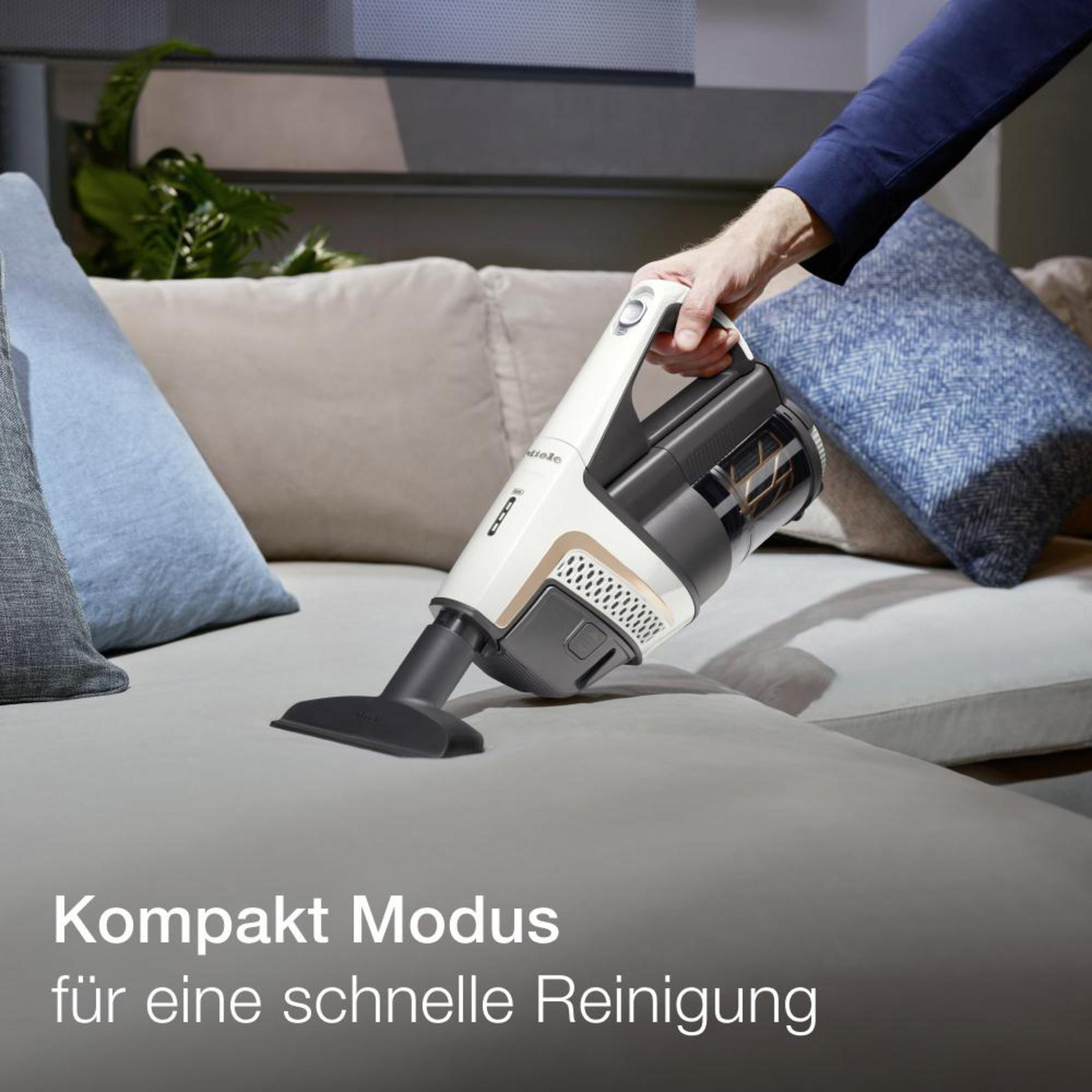 Ein Handstaubsauger saugt einen grauen Couch. Der Staubsauger ist weiß/schwarz. Kissen und Text im Hintergrund.