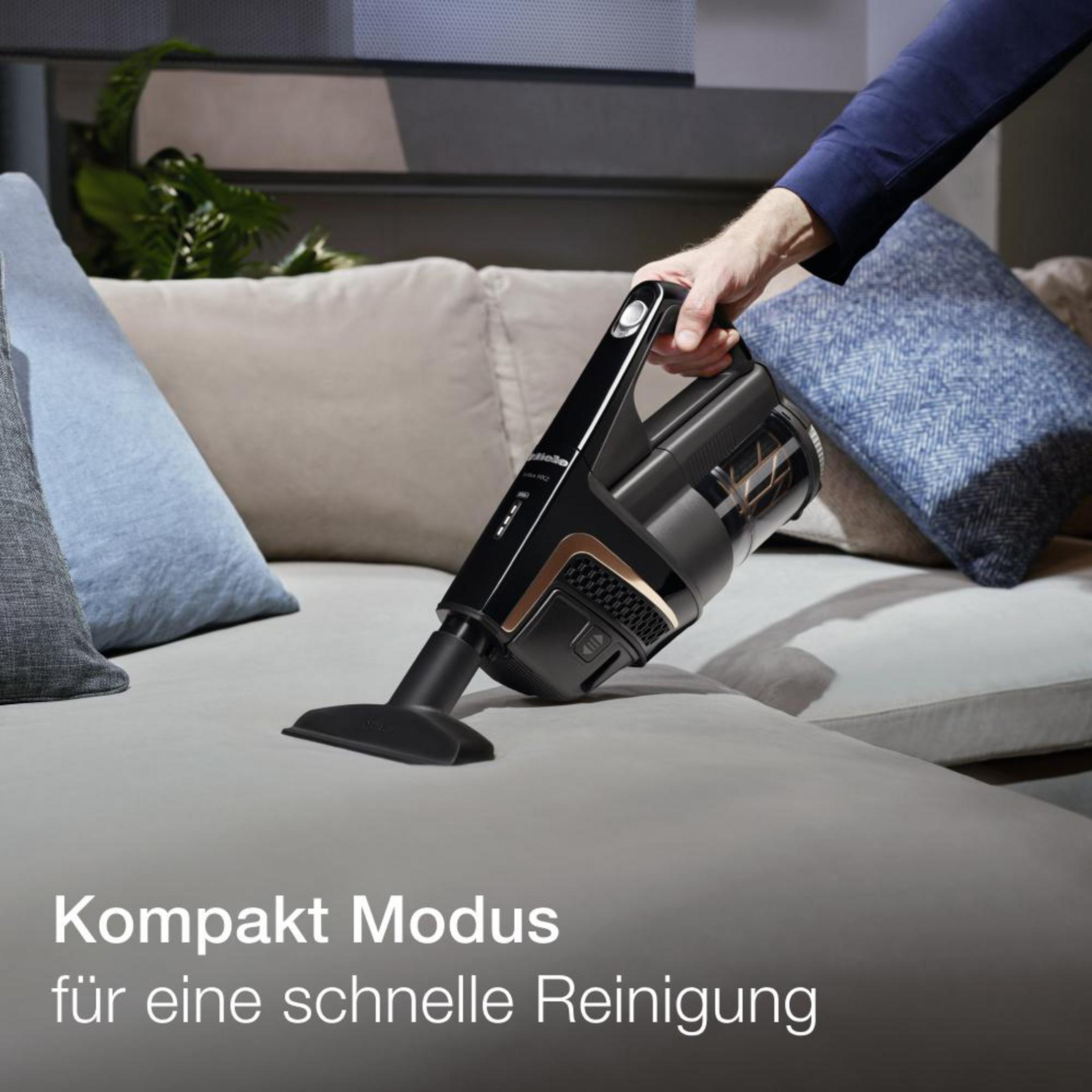 Eine Person benutzt einen schwarz-goldenen Staubsauger auf einem grauen Sofa. Der Kompaktmodus wird hervorgehoben.