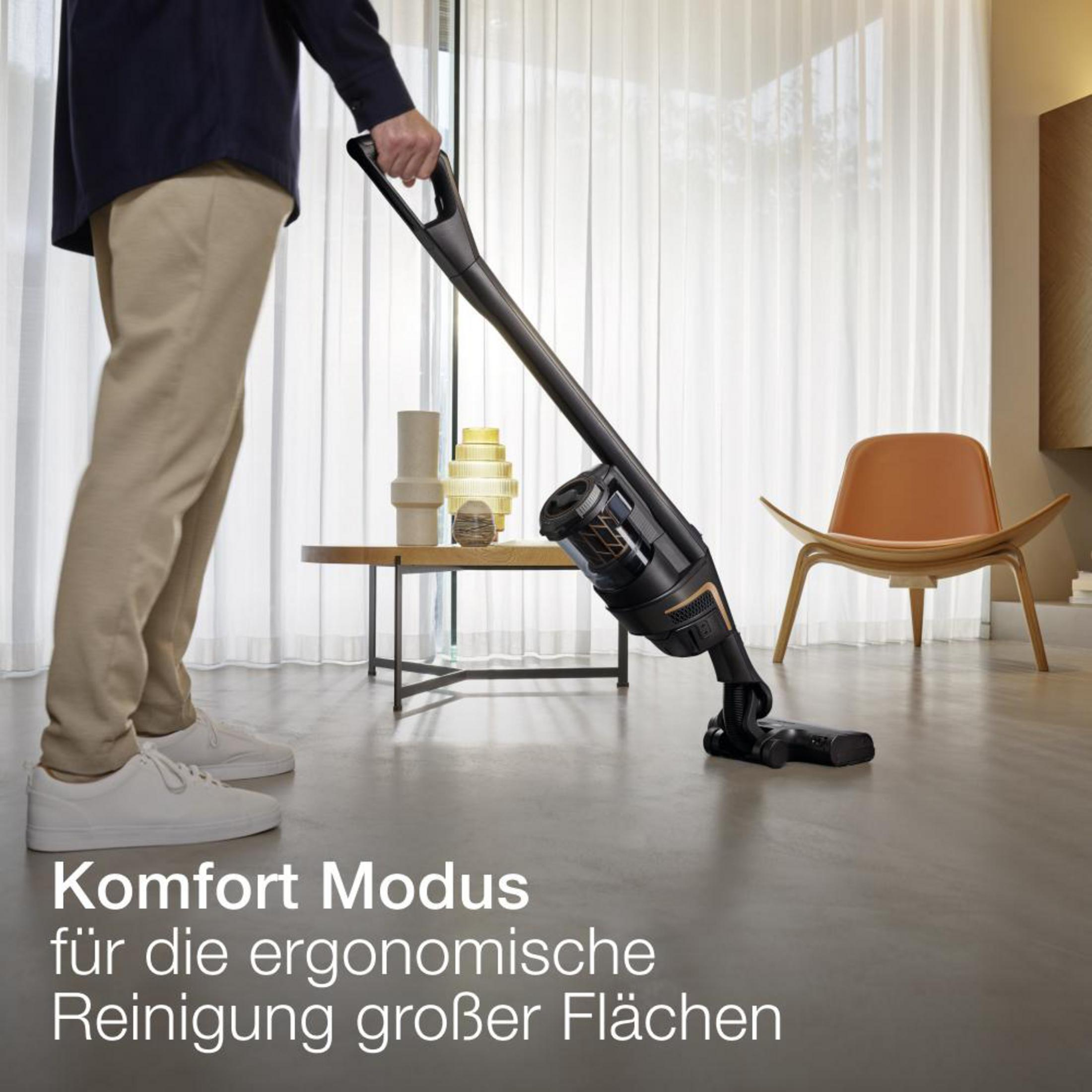 Eine Person, die mit einem schwarzen Akku-Staubsauger den Boden saugt, im Hintergrund Möbel. Deutscher Text.