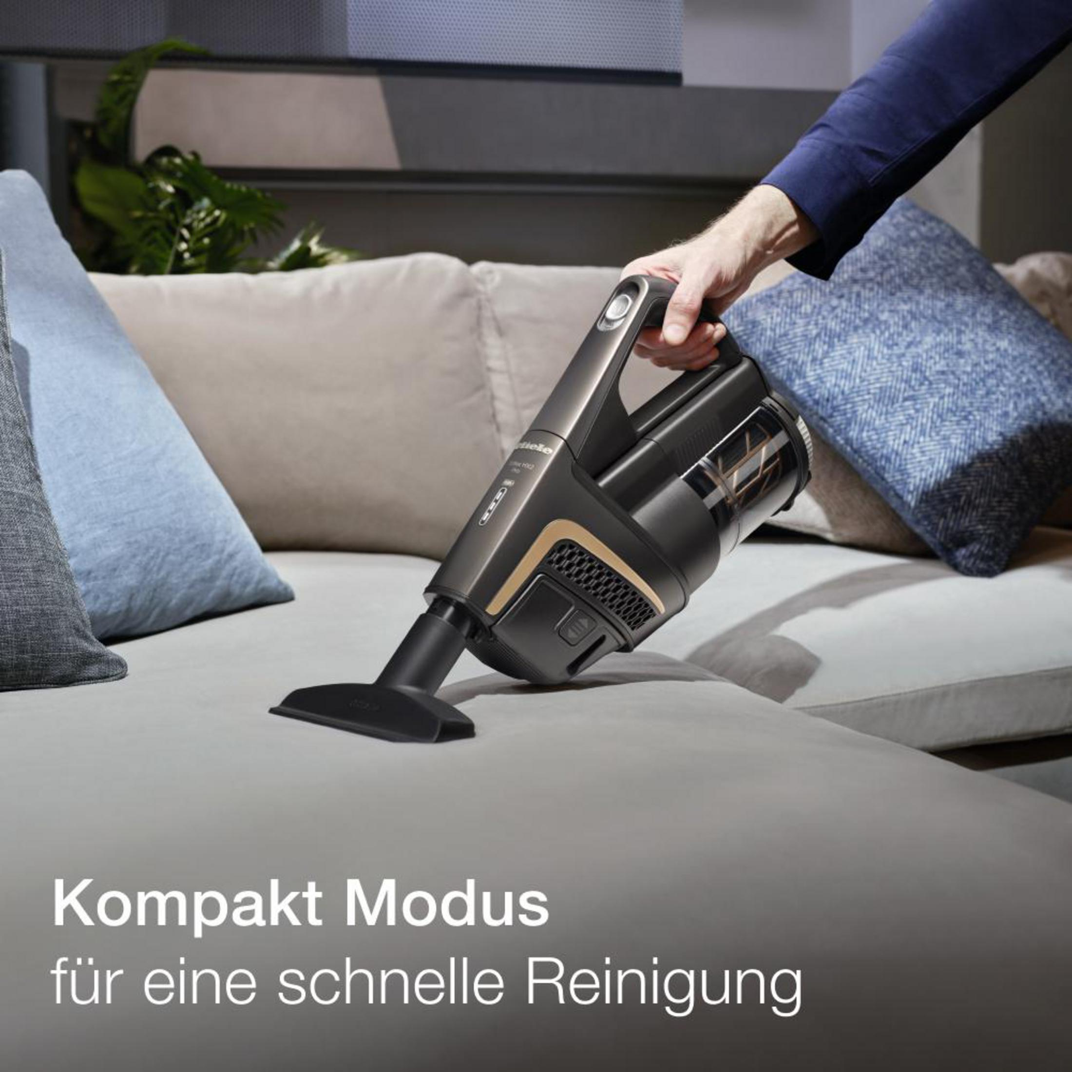 Hand hält einen schwarzen Staubsauger, der ein graues Sofa reinigt, Text im Kompaktmodus.