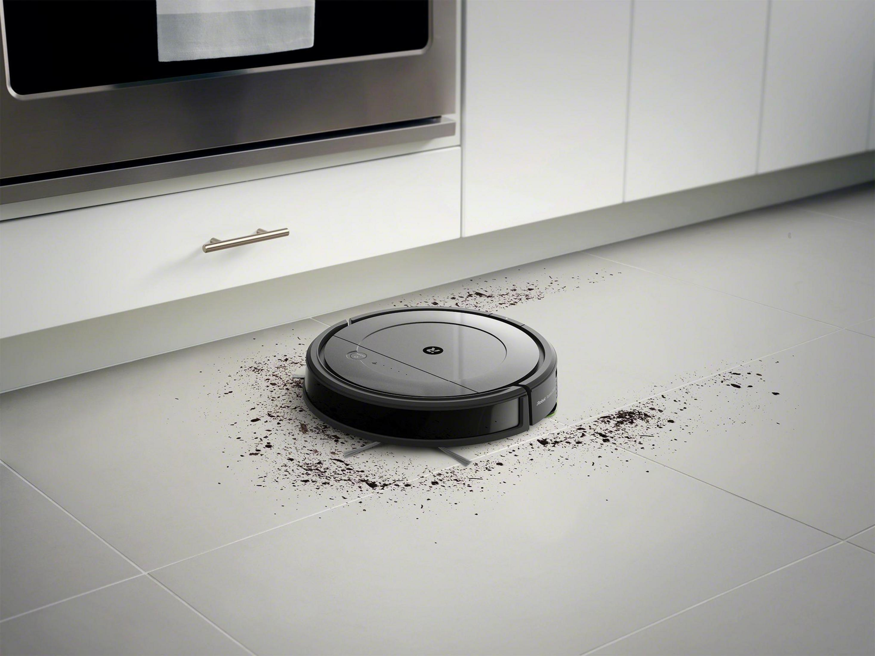 Robot aspirador negro en suelo baldosado blanco, limpiando restos. Armarios de cocina al fondo.