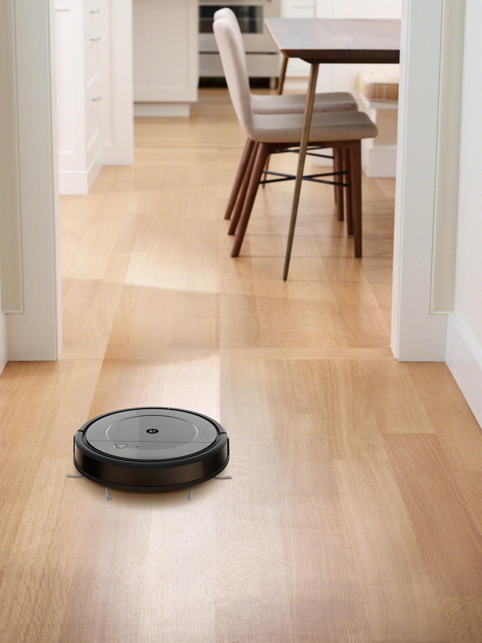 Una aspiradora robot negra en un piso de madera, en una habitación con una mesa y sillas visibles.