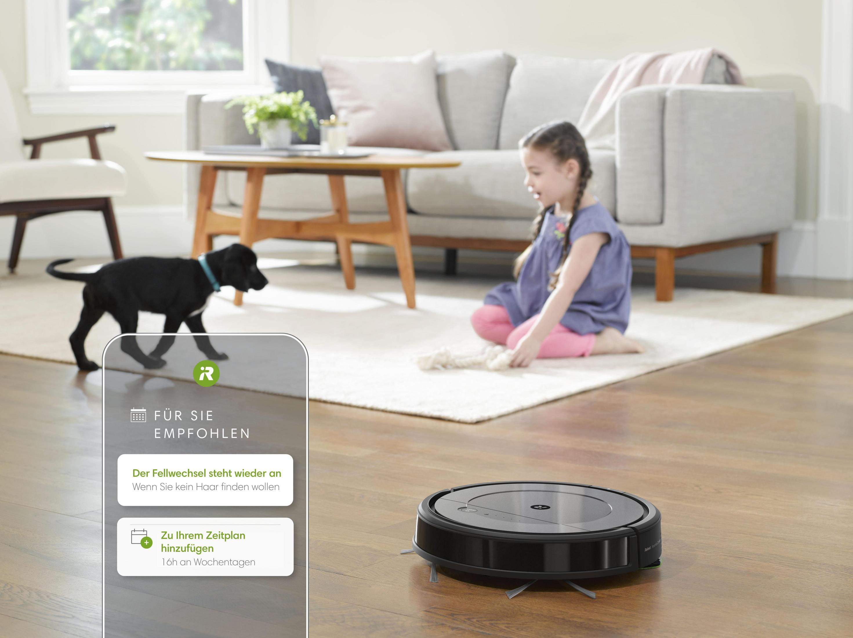 Niña, cachorro y aspiradora robot en piso de madera, ambiente de sala.