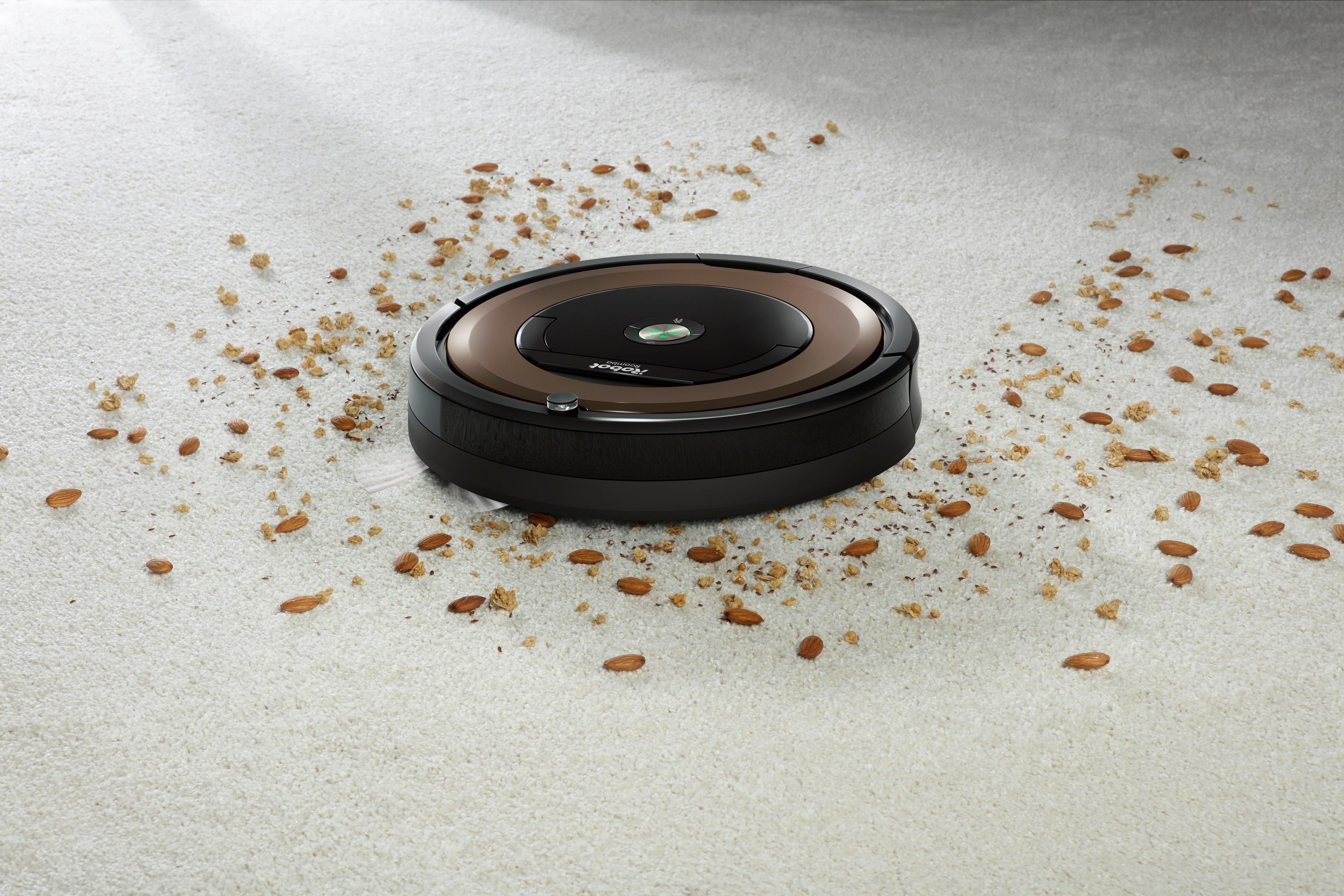 Un aspirapolvere robotico rotondo, marrone e nero su un tappeto bianco con mandorle sparse.
