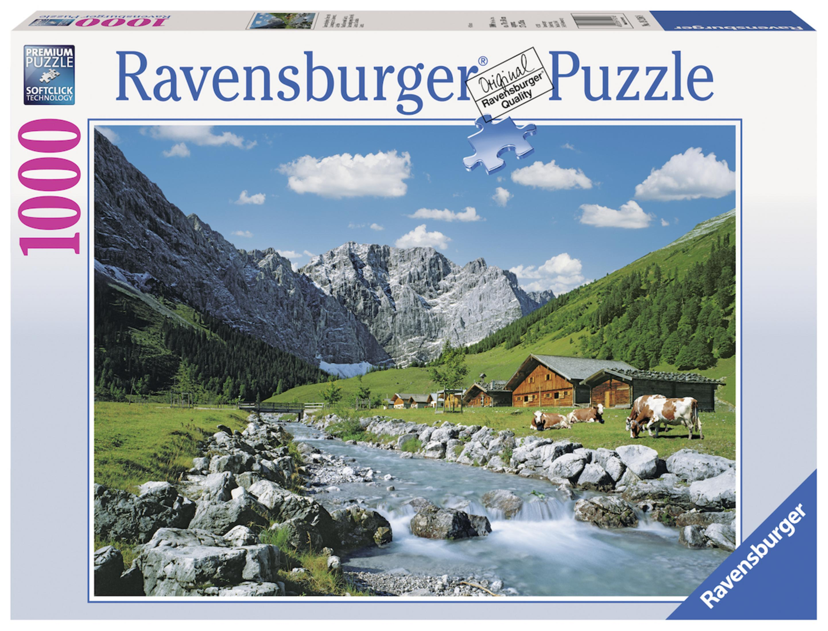 Pudełko puzzli Ravensburger. Krajobraz górski i rzeki z pasącymi się krowami. Błękitne niebo z chmurami.