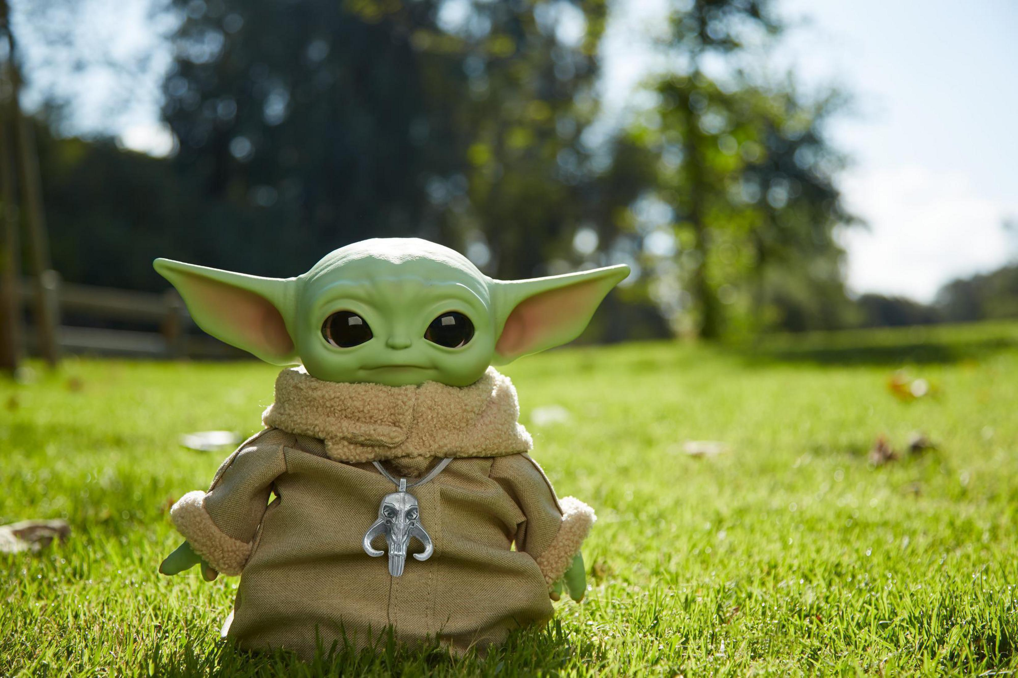 Lalka Baby Yoda na trawiastym polu. Nosi swój płaszcz i naszyjnik. Tło jest rozmyte.