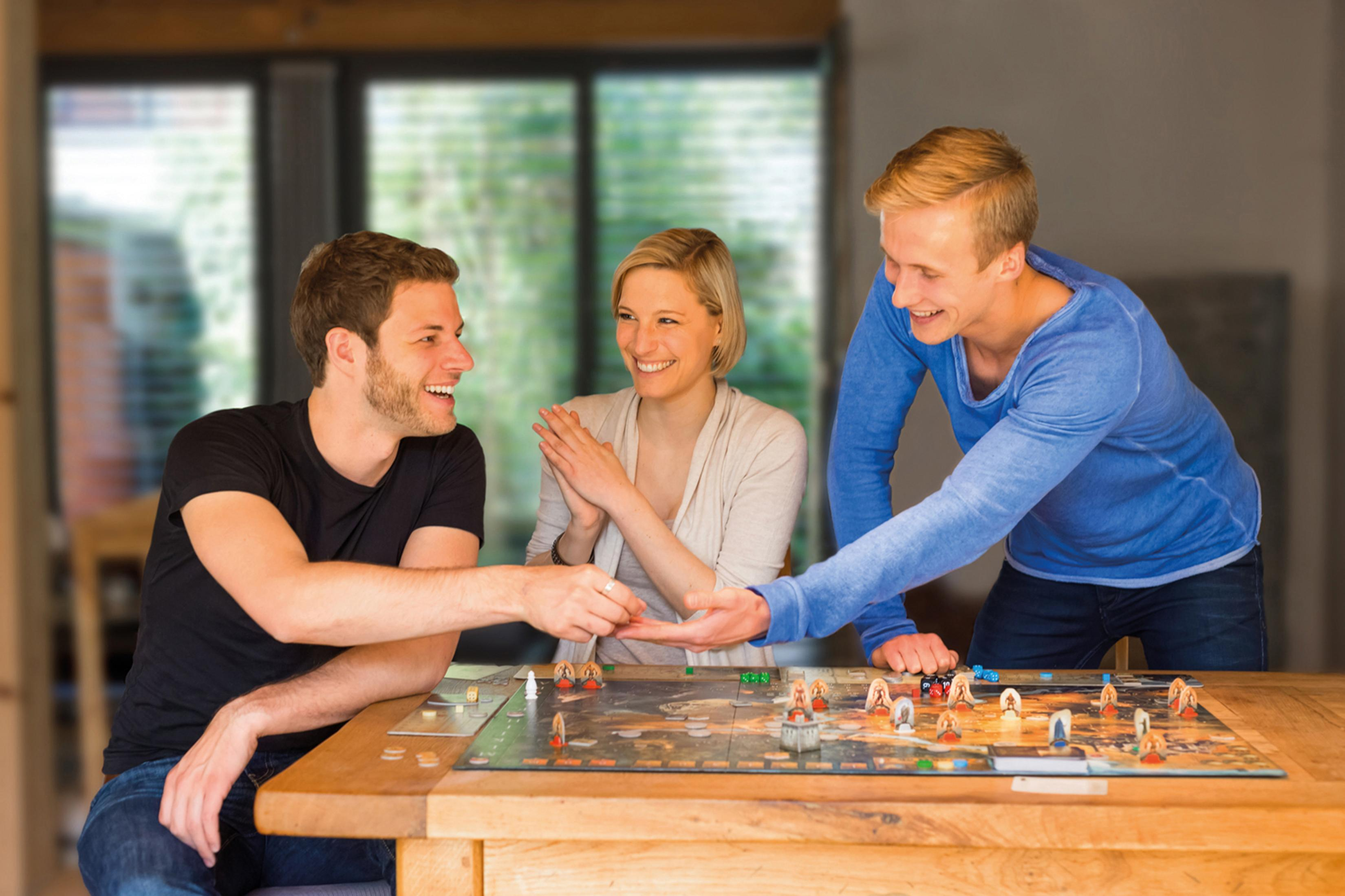 Drei Personen spielen ein Brettspiel an einem Tisch. Sie lächeln.